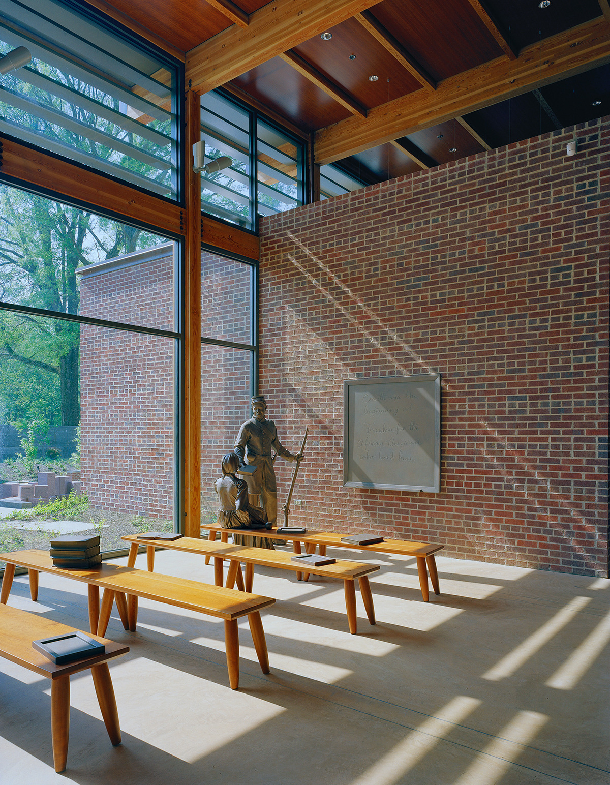 A sunlit room with brick walls, wooden benches, and a large window. In this Corinth Civil War interpretive center, a statue of a seated figure holding a staff is positioned next to a wall plaque.