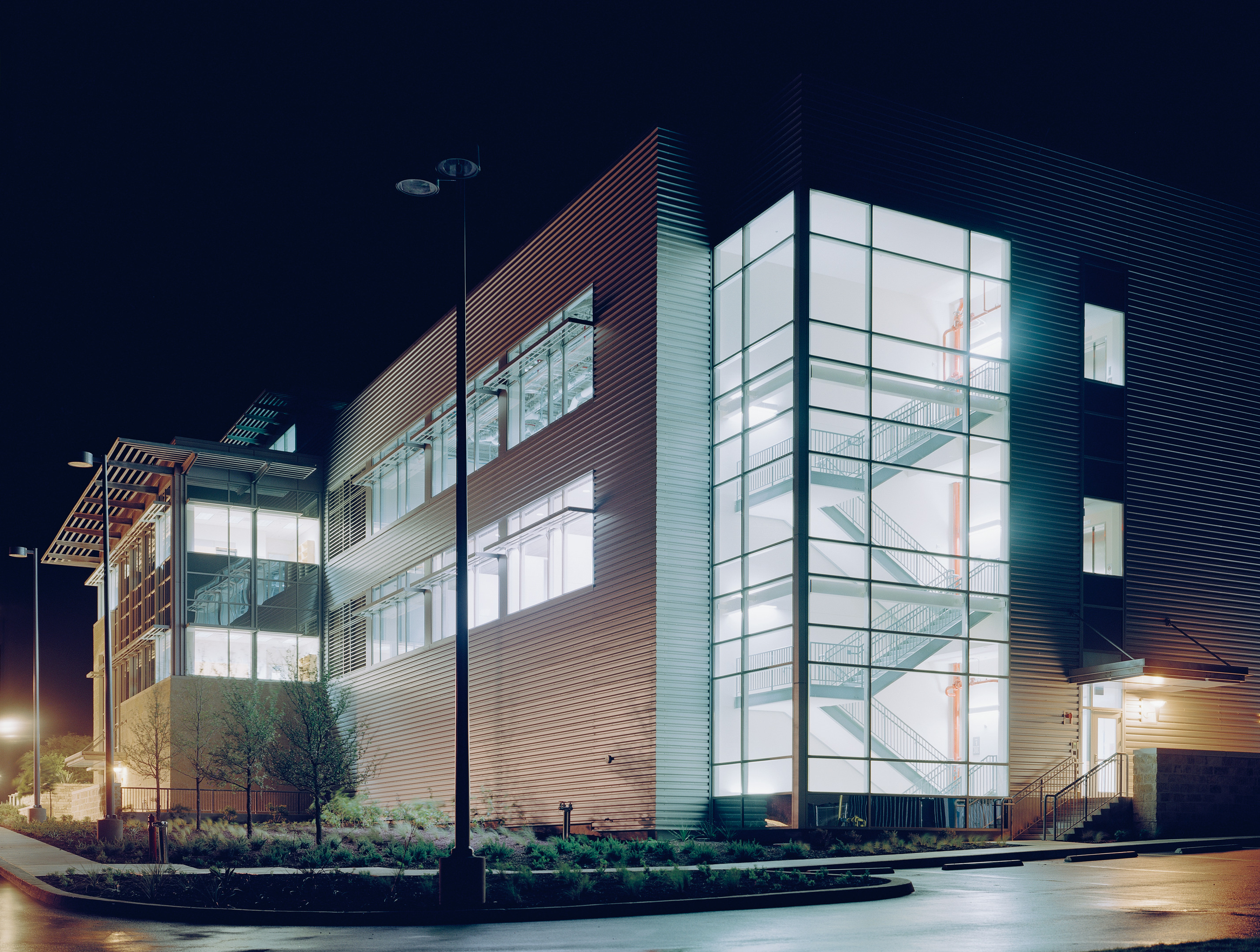 A modern, illuminated office building at night featuring large glass windows, multiple floors, visible staircases, and a landscaped exterior creates an auto draft of elegance and sophistication.