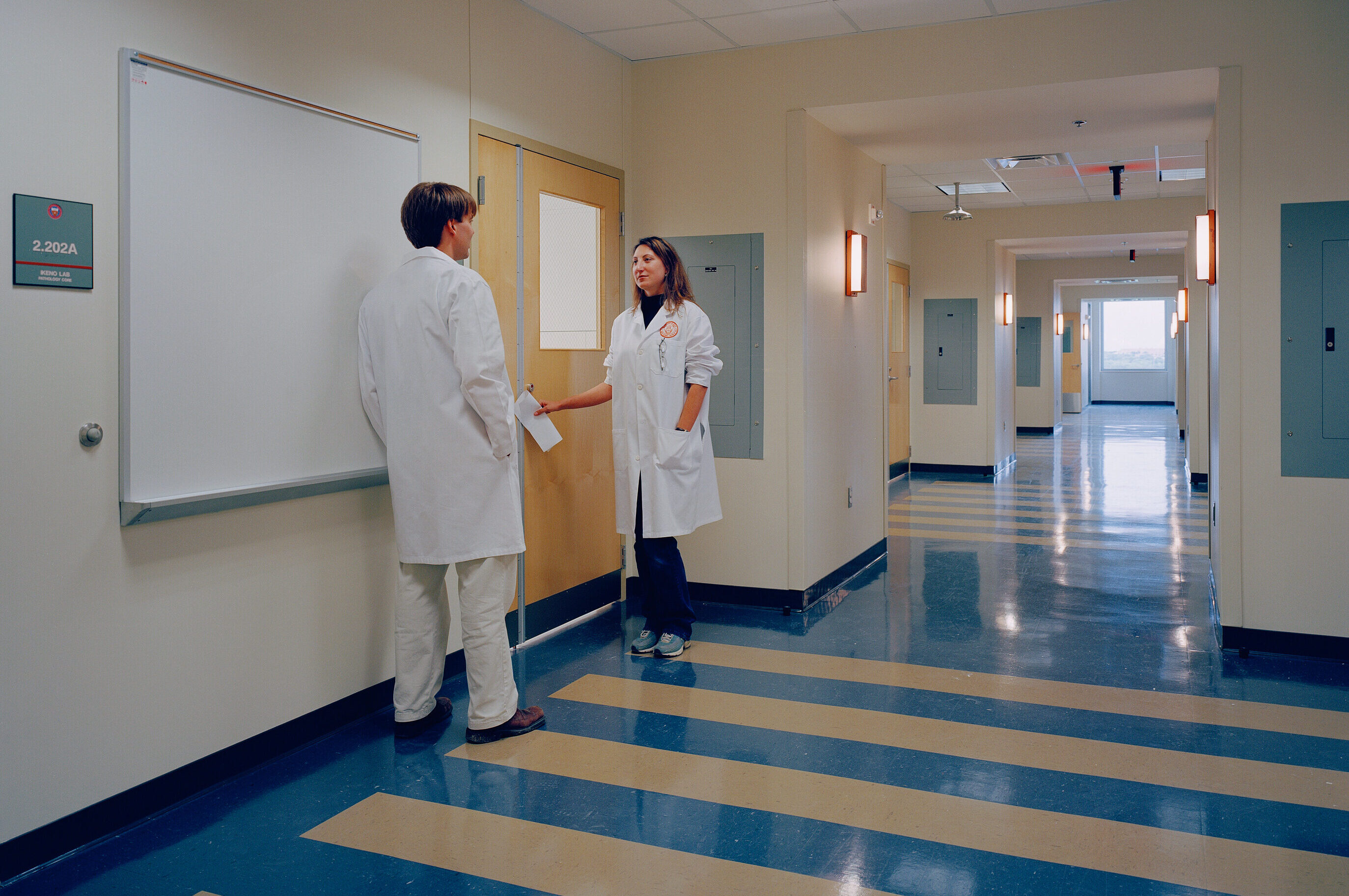 Two medical professionals in white lab coats confer in a well-lit hospital hallway with blue and white flooring, discussing an auto draft update for patient records.