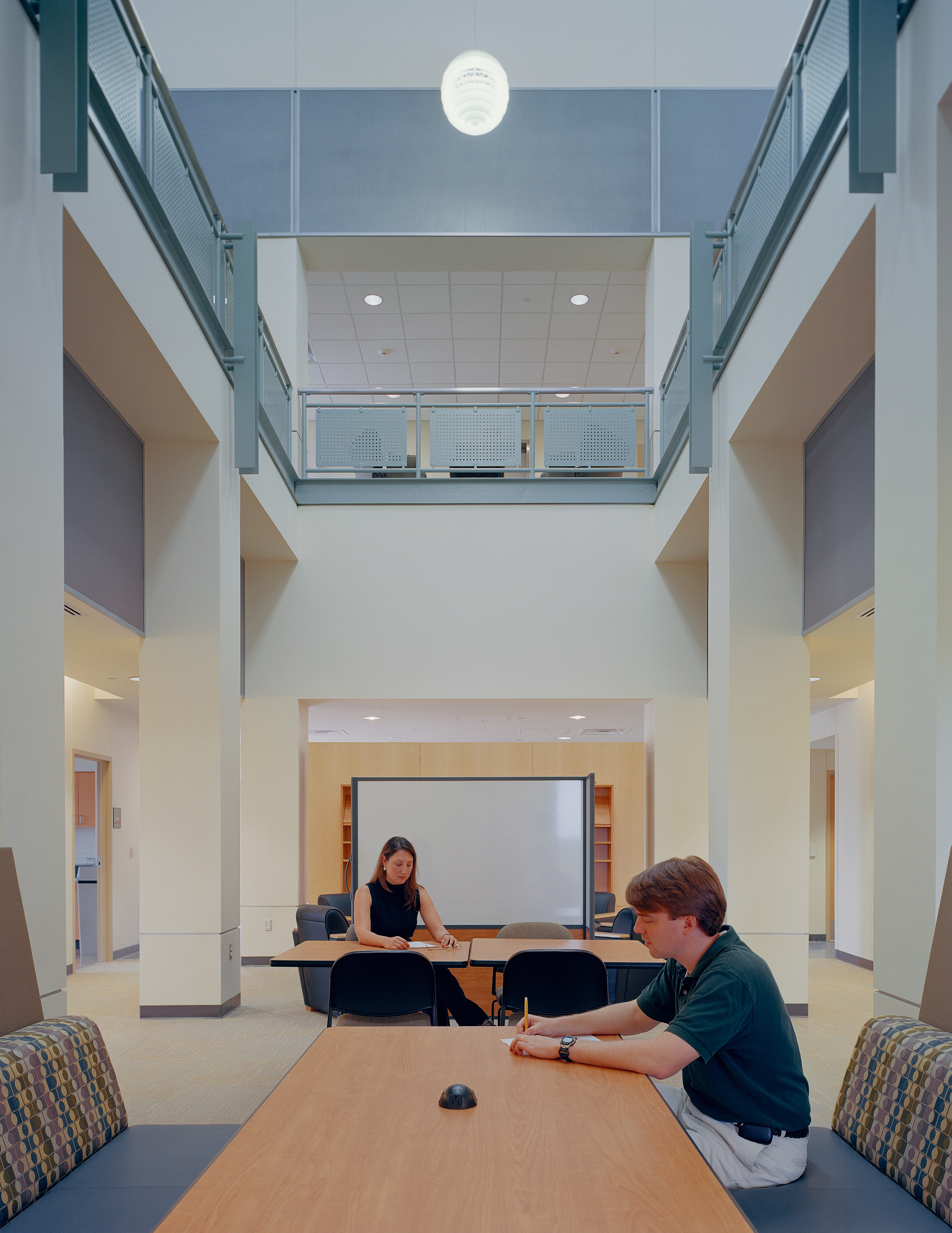 Two people in a modern, high-ceilinged study area with tables and chairs. One person is writing at a table while the other leisurely engages in an Auto Draft, seated further back.