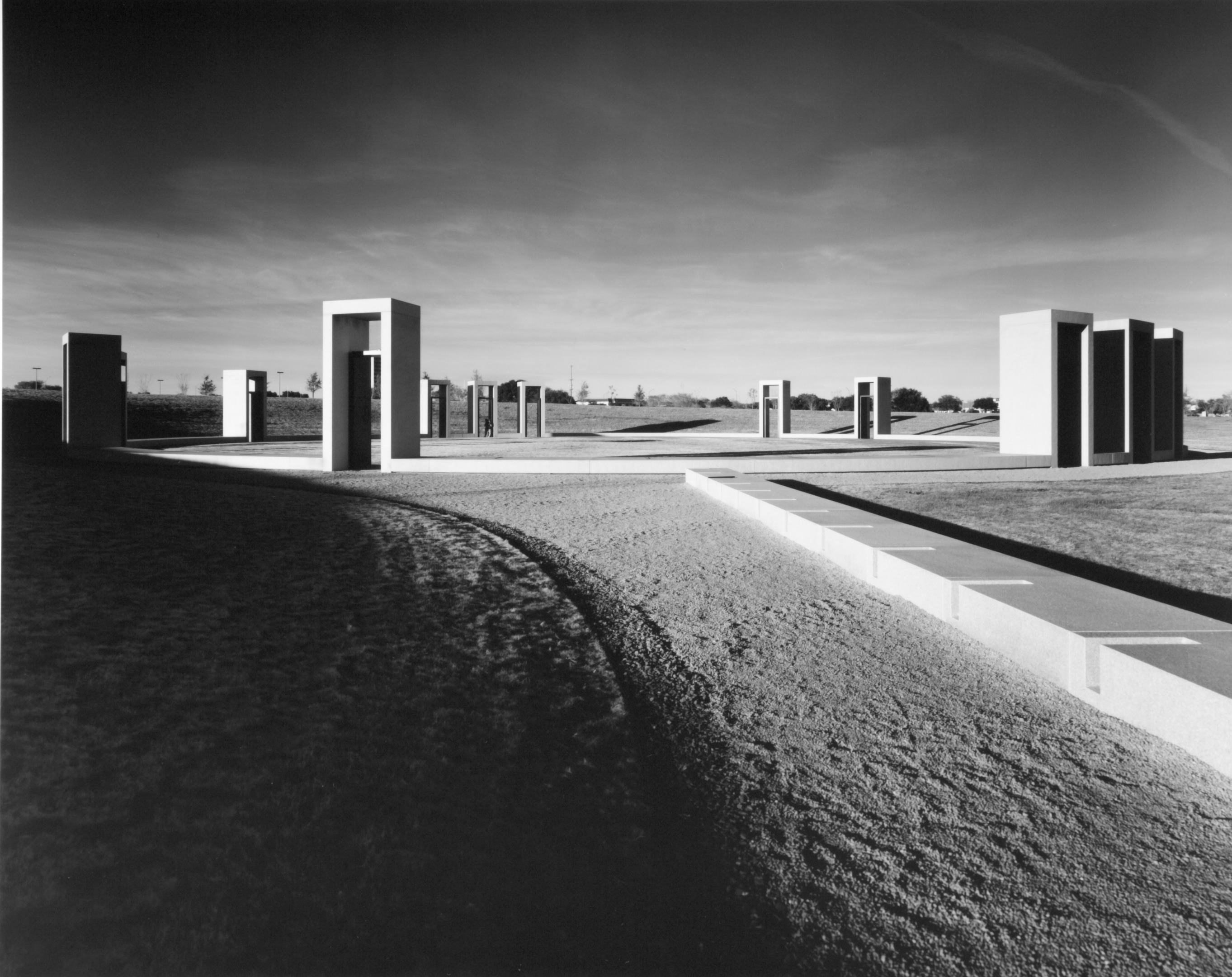 A black-and-white photo of the Bonfire Memorial at Texas A&M University, featuring multiple rectangular stone structures arranged in a geometric pattern under a cloudy sky.