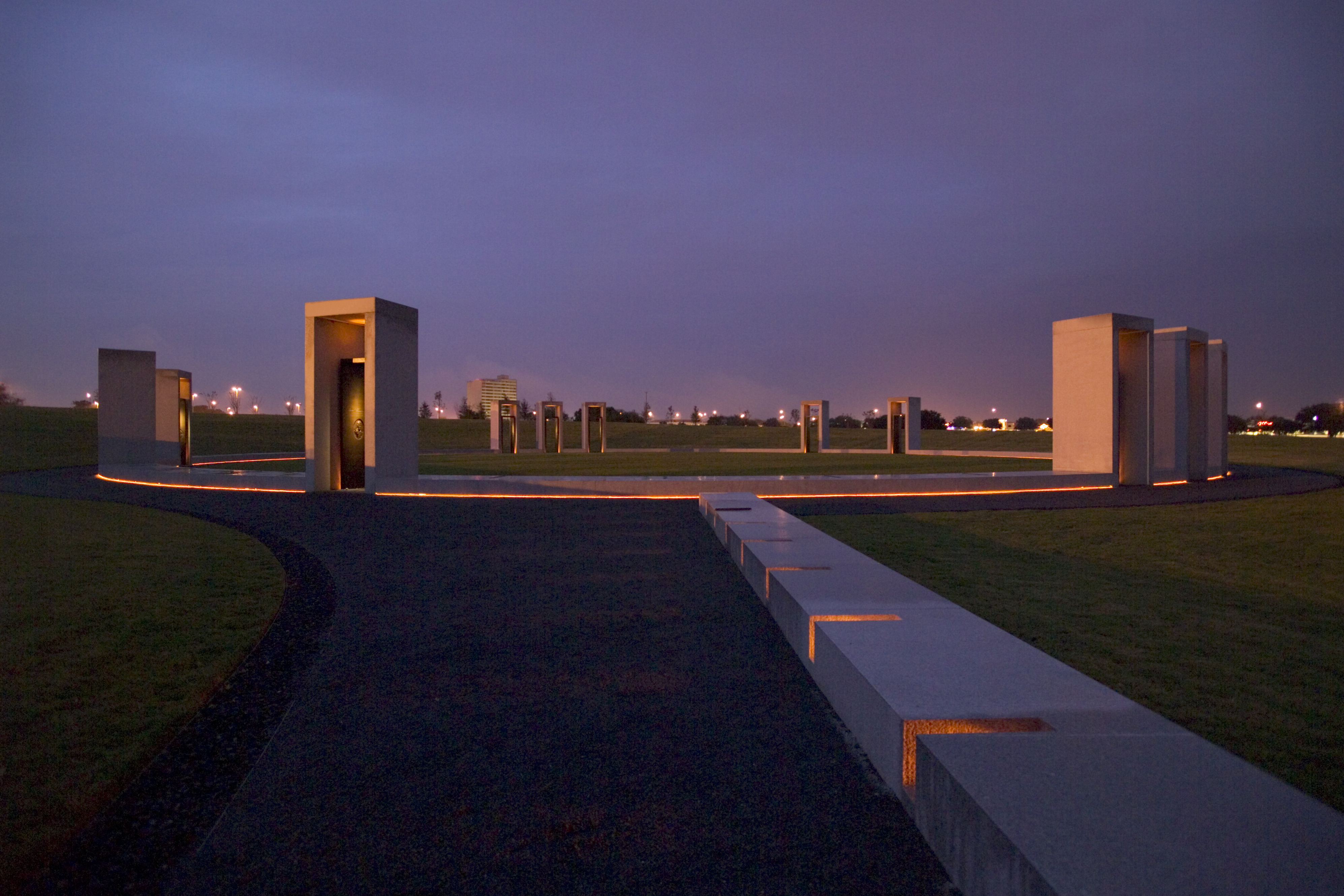 A twilight view of a modern outdoor monument with illuminated pathways, vertical stone structures, and an open grassy area. Urban buildings and lights are visible in the background.