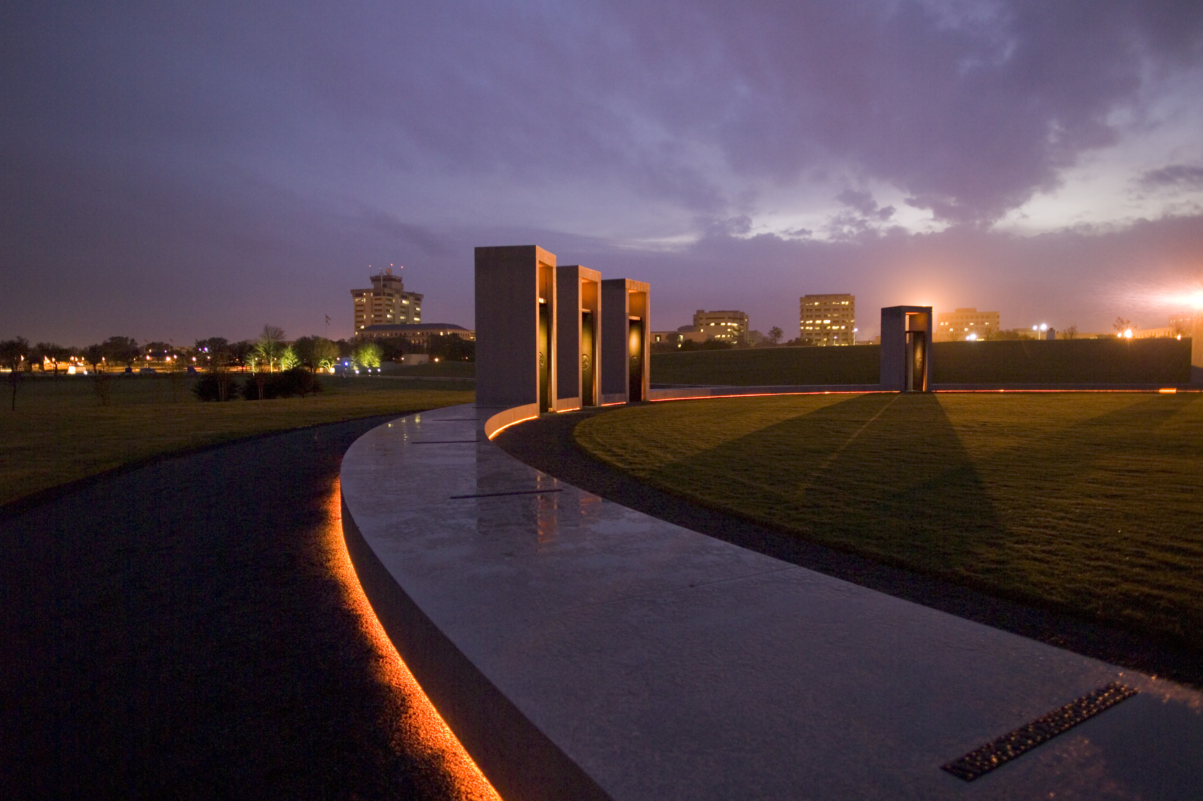 Twilight view of a monument featuring tall stone structures and a curved pathway, with lights illuminating the scene against a backdrop of city buildings and a darkening sky.