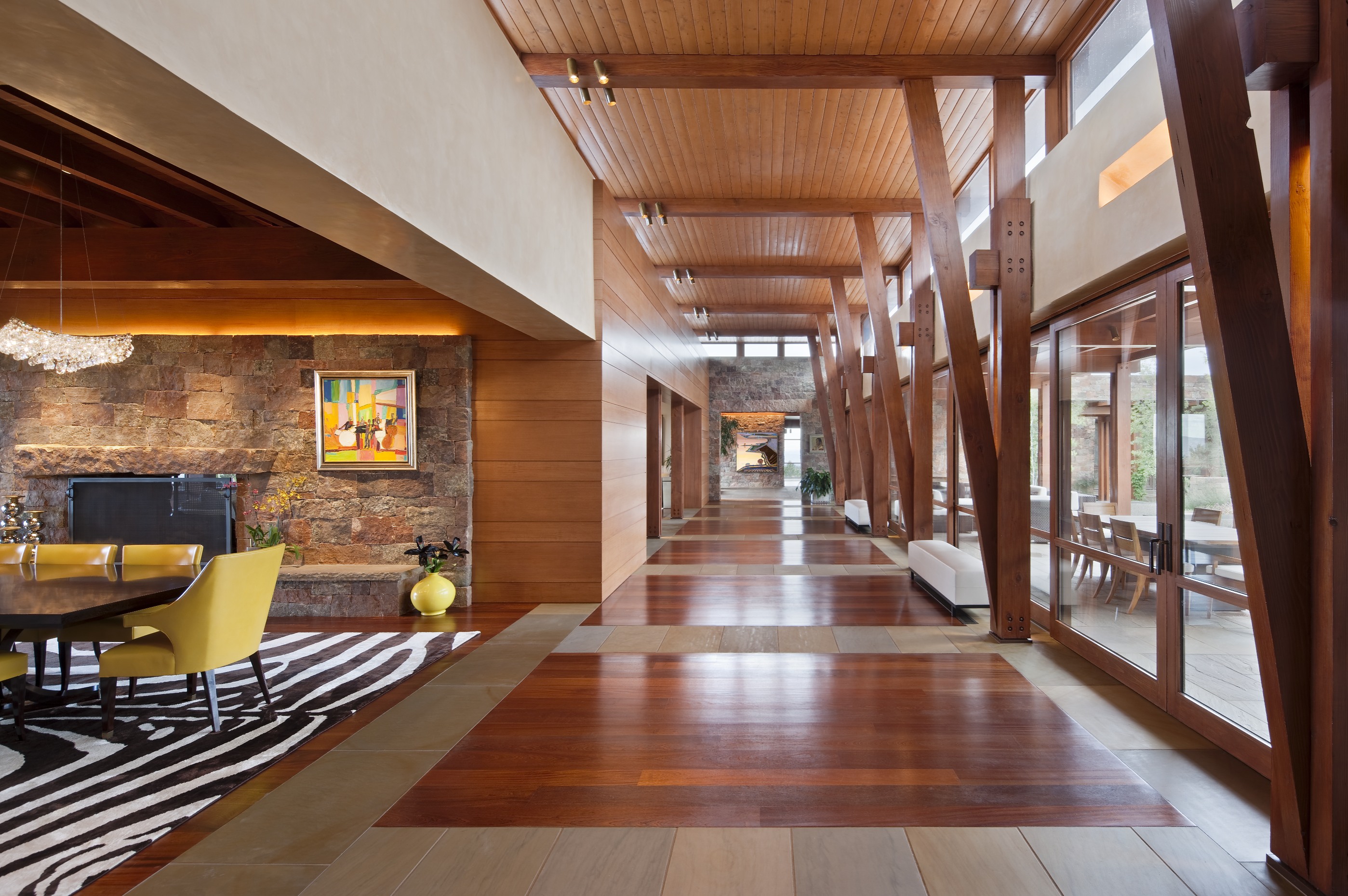 A spacious, modern hallway with wooden beams and paneling evokes a Santa Fe retreat, leading to a brightly lit dining area adorned with art and a zebra-striped rug. A large window wall provides ample natural light.