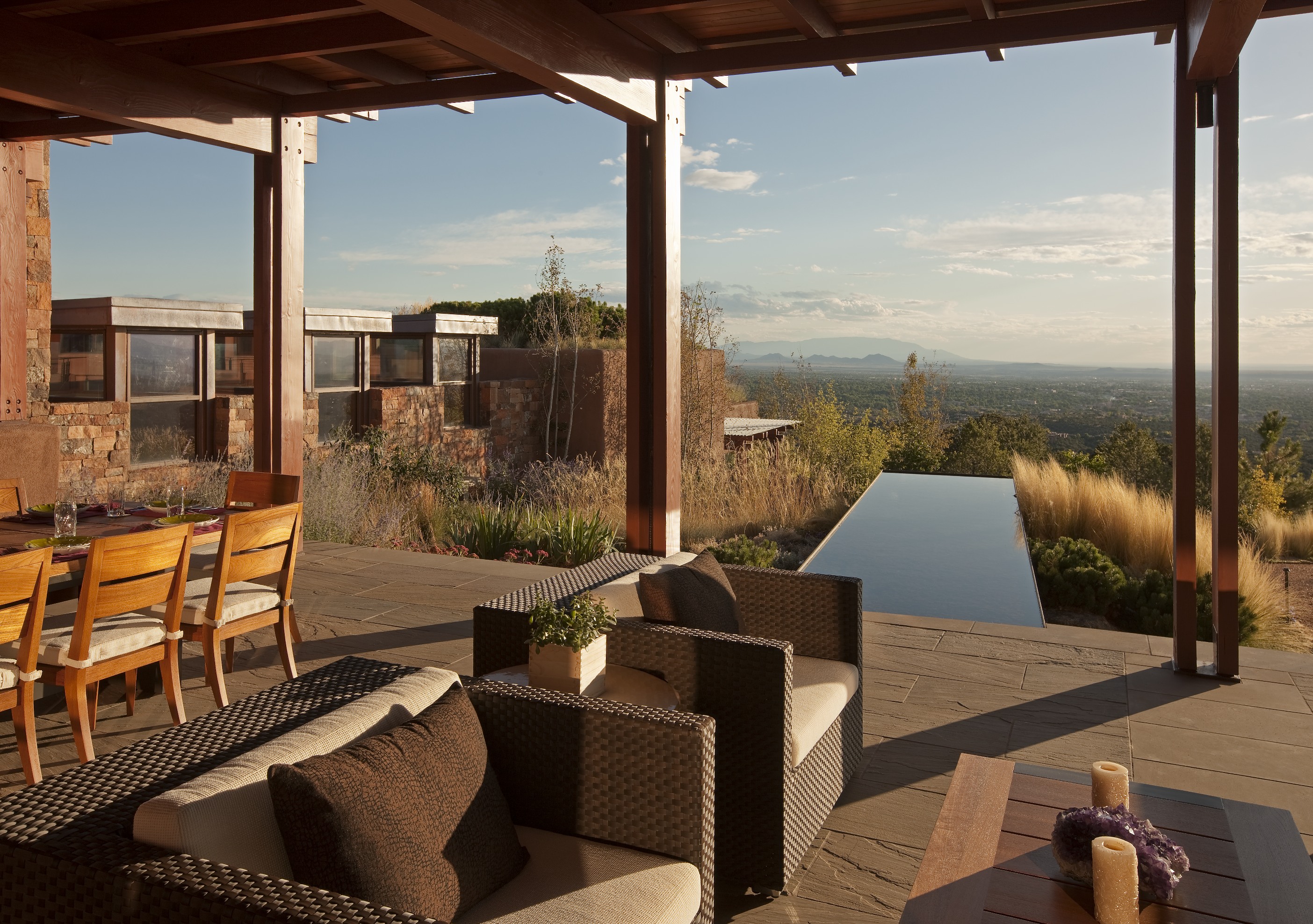 Outdoor patio area with wicker furniture, a wooden dining table, and a view of a modern Santa Fe home with an infinity pool overlooking a vast landscape in the background, creating the perfect retreat.