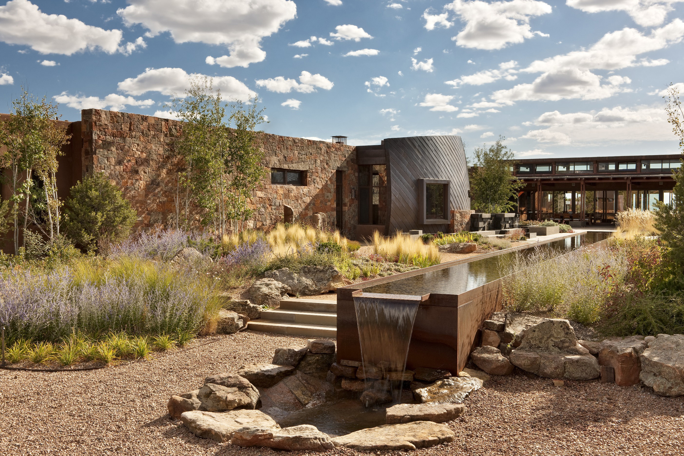 A modern retreat with stone walls and metal accents, surrounded by landscaped gardens, evokes the charm of Santa Fe. A water feature runs through the garden under a partly cloudy sky.