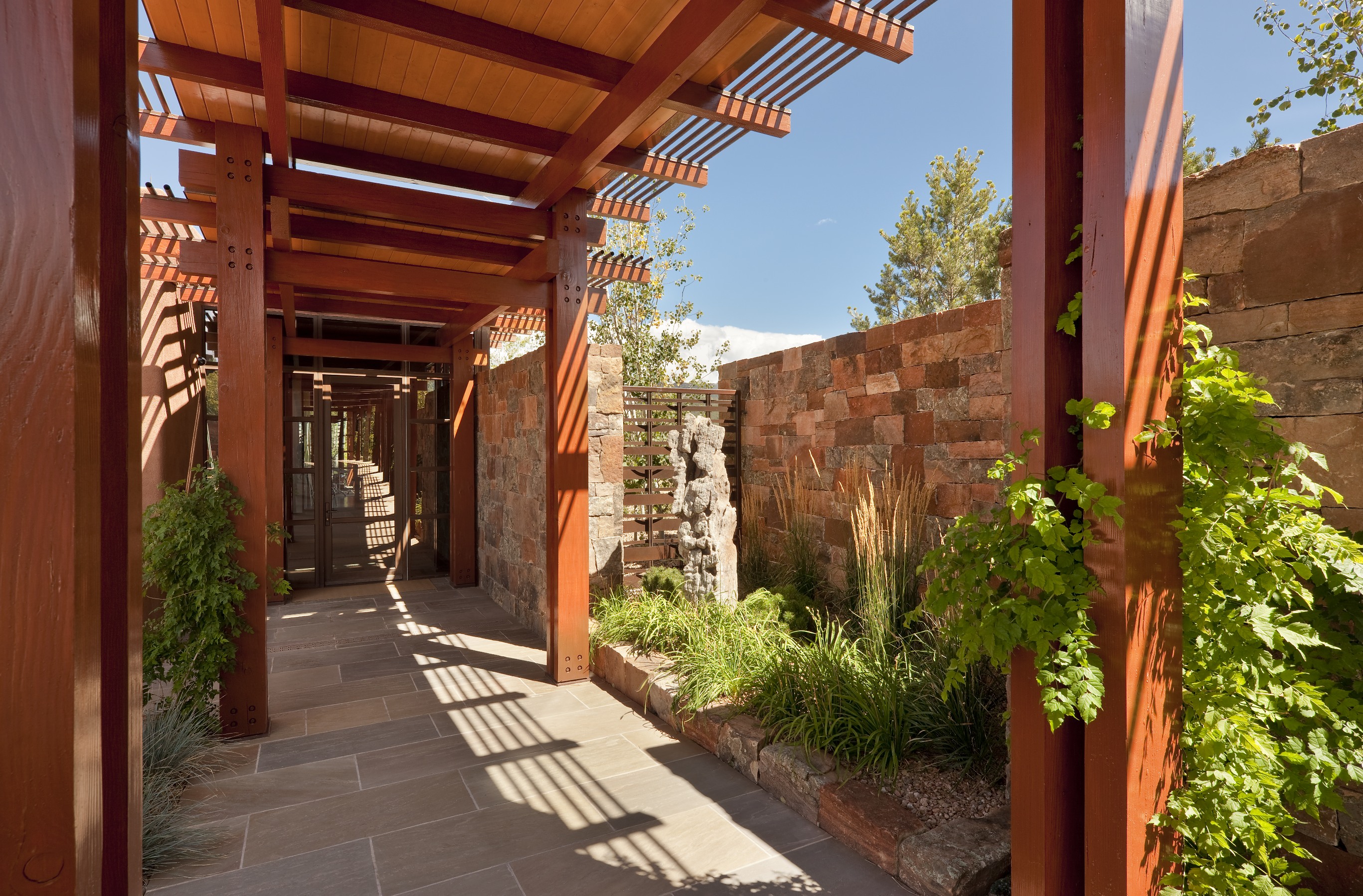 A walkway with a wooden pergola and stone walls, featuring climbing plants and sunlight casting shadows on the ground, creates a serene Santa Fe retreat.