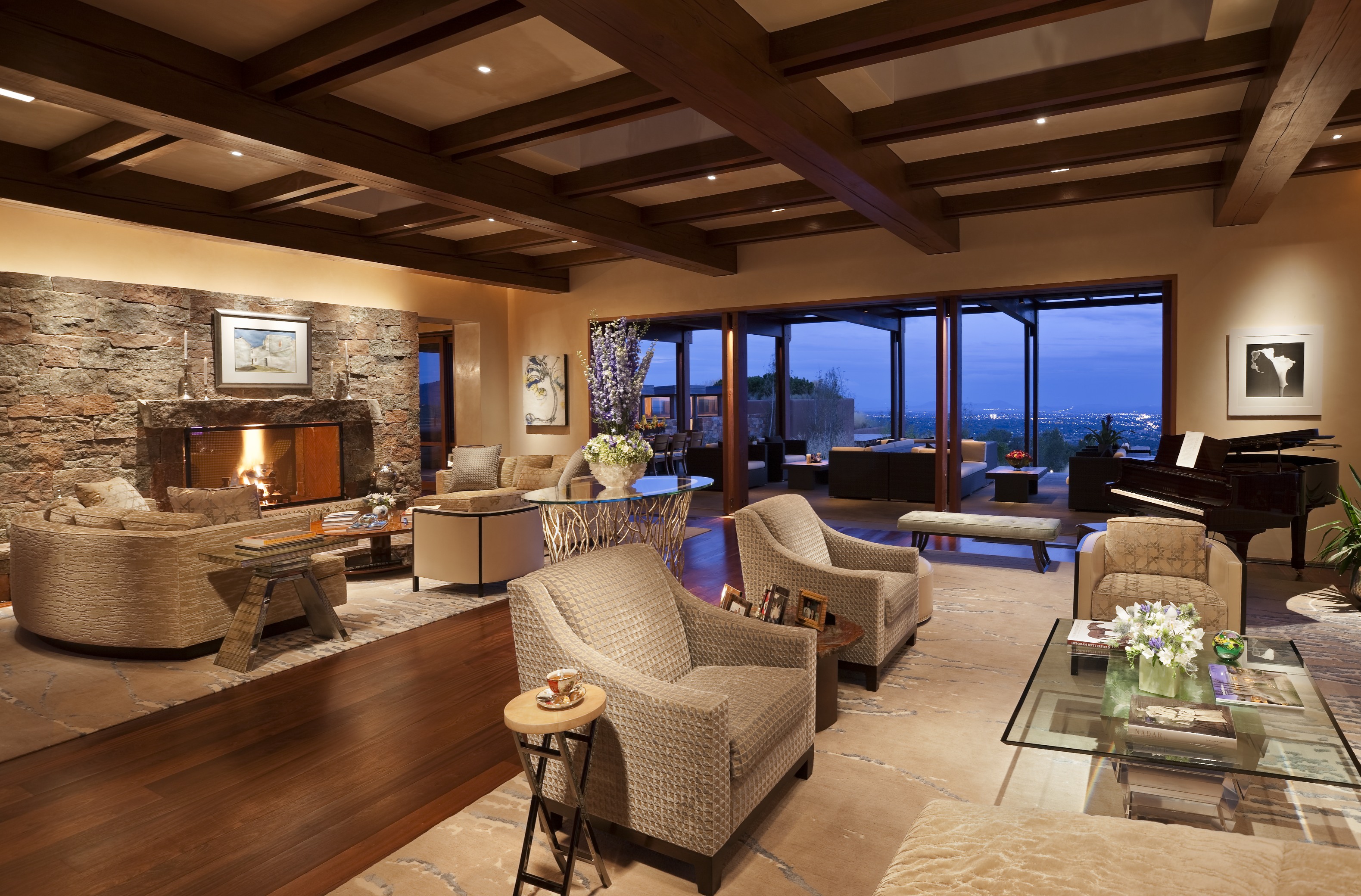 A spacious living room with a stone fireplace, wooden beams, and modern furniture. Large glass doors open to a patio with a view of the city lights at dusk, creating the perfect Santa Fe retreat ambiance.