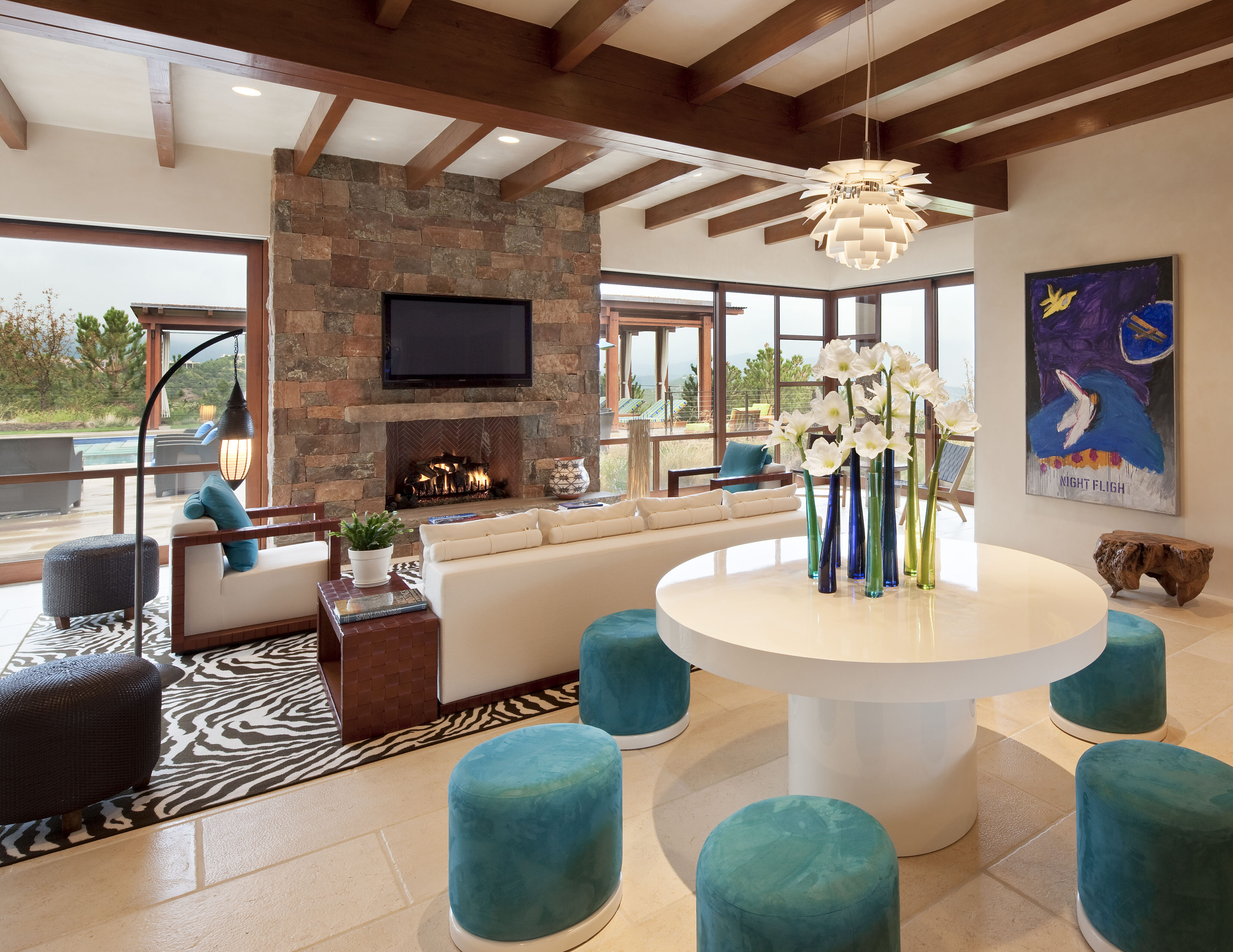 Modern living room with a stone fireplace, wall-mounted TV, white sofa, round table with flower vase, blue stools, and abstract art. Large windows reveal an outdoor seating area and scenic view reminiscent of a Santa Fe retreat.