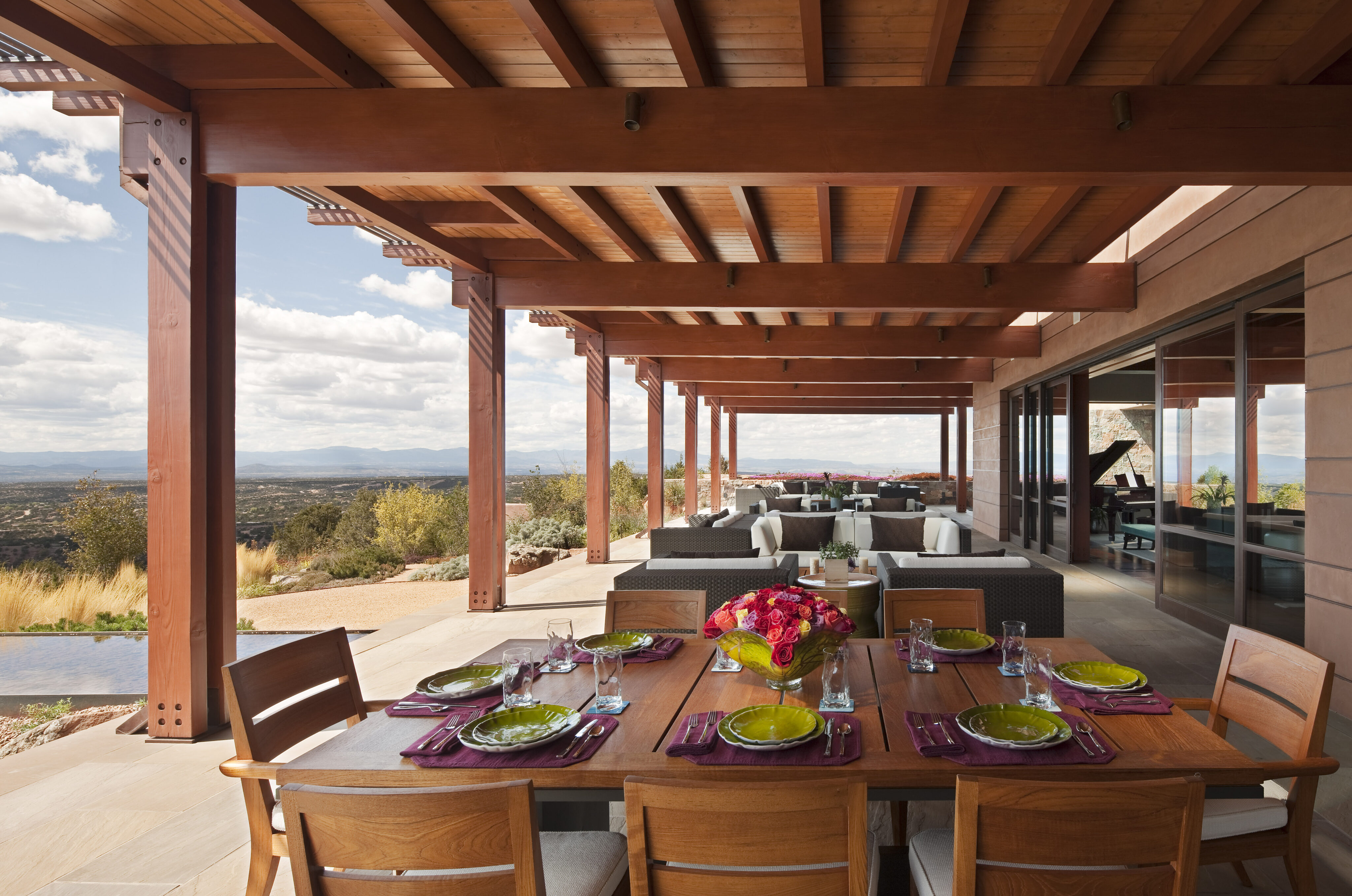 An outdoor patio at this Santa Fe retreat features a wooden dining table set with dishes and glasses, surrounded by chairs. Lounge seating is visible in the background under a wooden pergola with scenic views beyond.