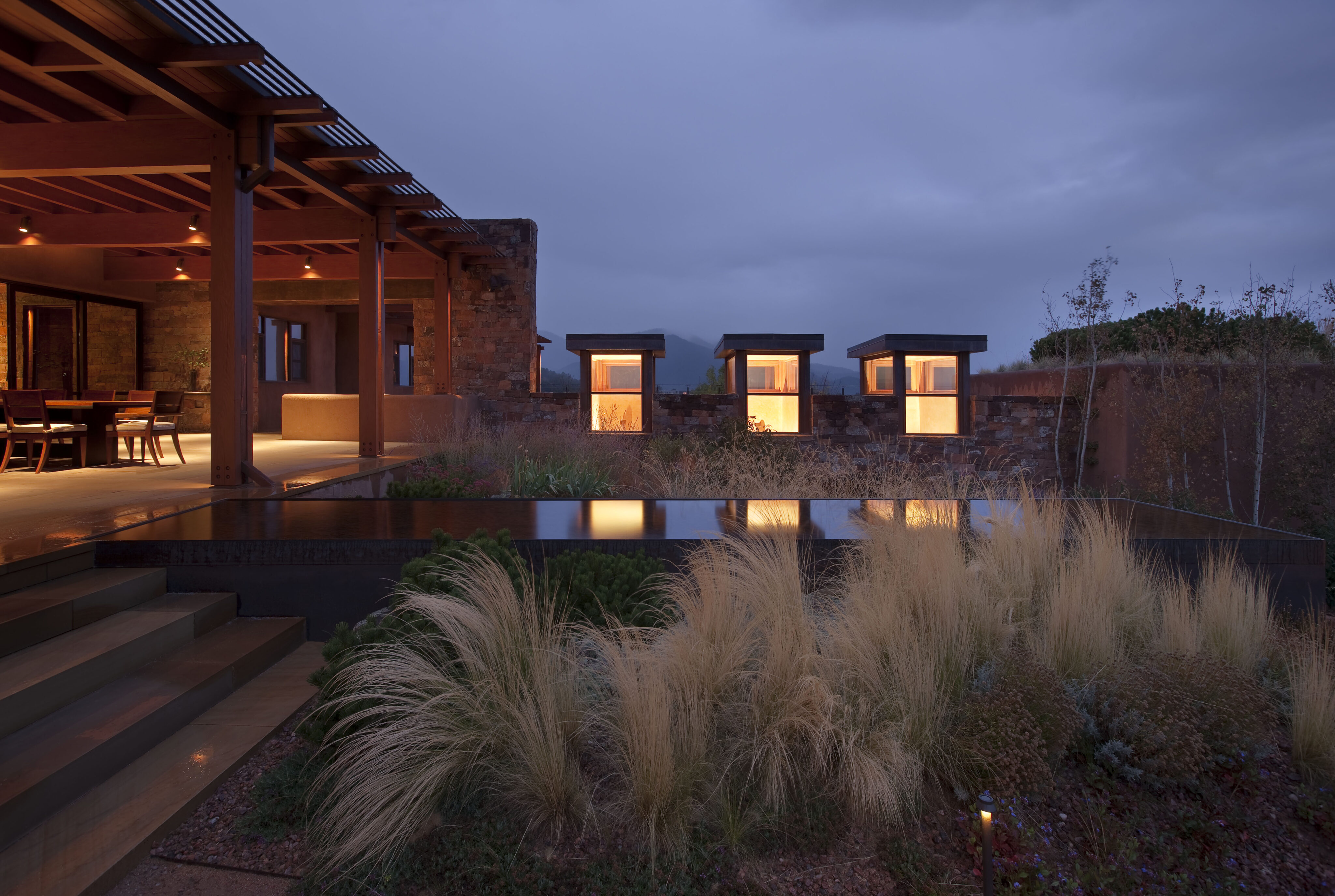 A modern Santa Fe retreat at dusk, featuring an outdoor patio with warm interior lighting and a reflective pool in the foreground, surrounded by ornamental grass.