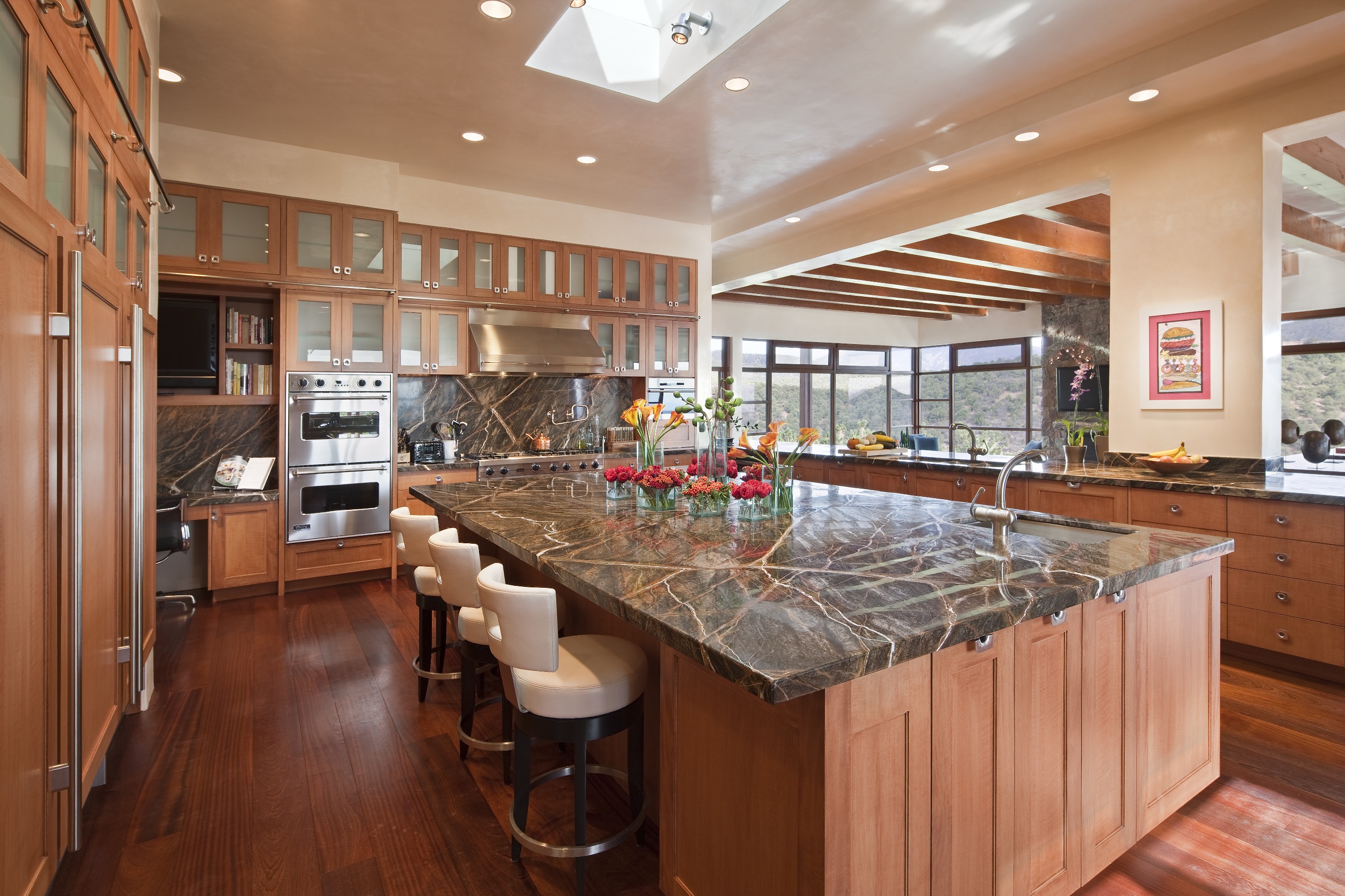 Modern kitchen with wooden cabinetry, a large marble island with bar stools, stainless steel appliances, and hardwood flooring. Sunlight streams through a skylight and large windows, illuminating the space. This Santa Fe-inspired retreat seamlessly blends comfort and elegance.