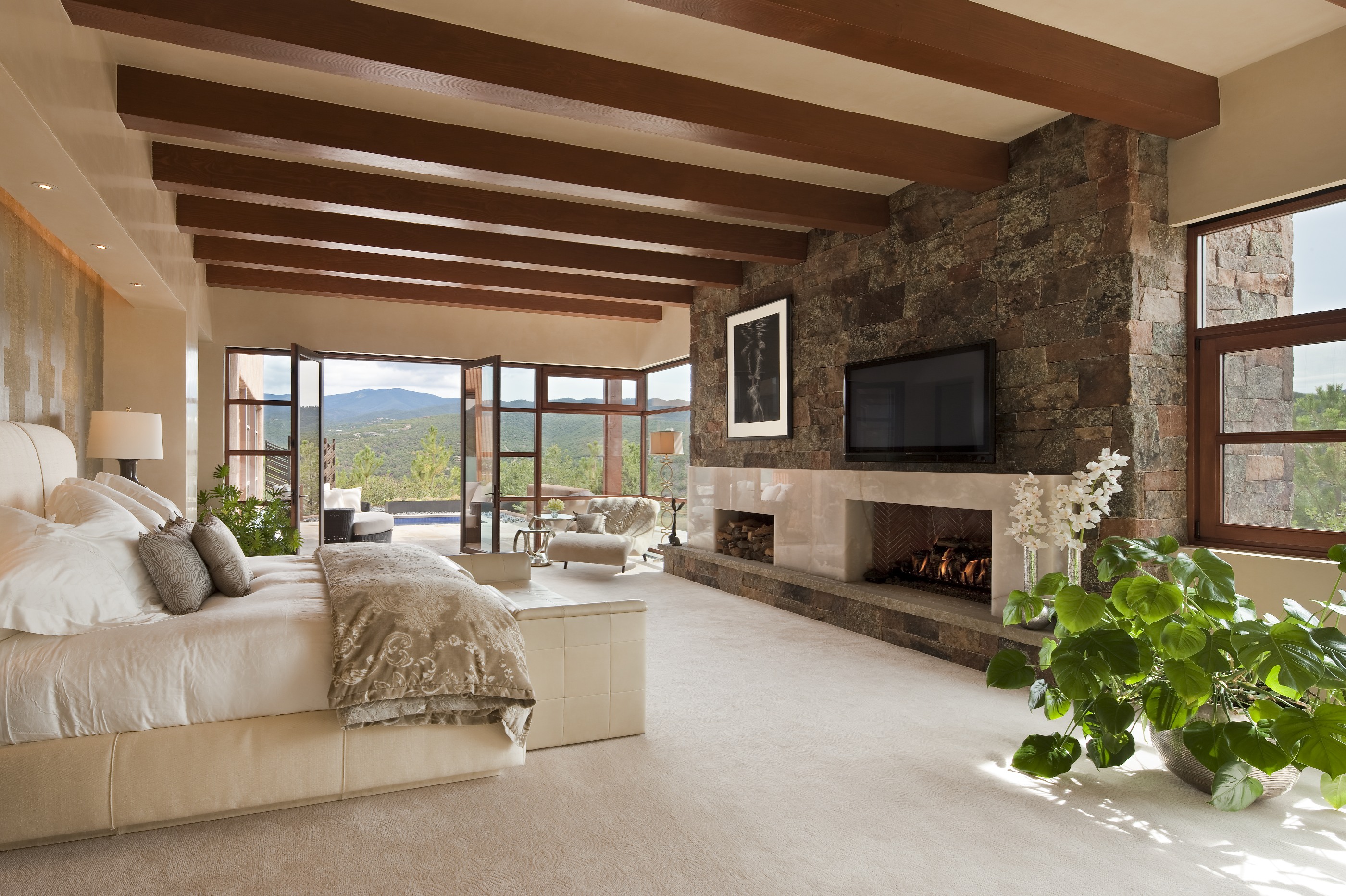 A spacious Santa Fe retreat featuring exposed wooden beams, a large bed, stone fireplace, television, and large windows offering a scenic mountain view. The room also boasts lush indoor plants and a light color scheme.