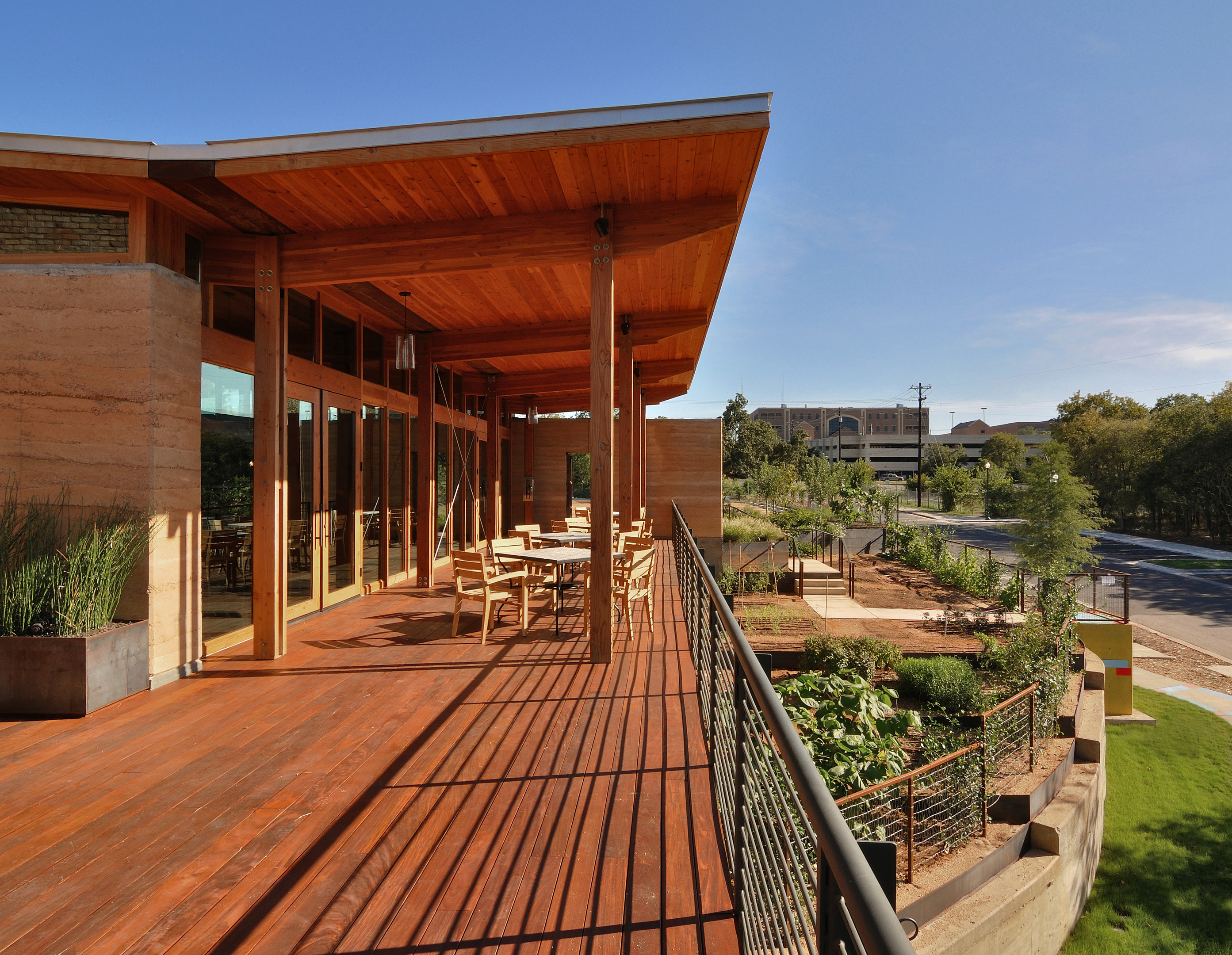 Wooden deck with tables and chairs attached to a modern building, overlooking a landscaped garden, and bustling street—a perfect spot for enjoying an auto draft of city life.