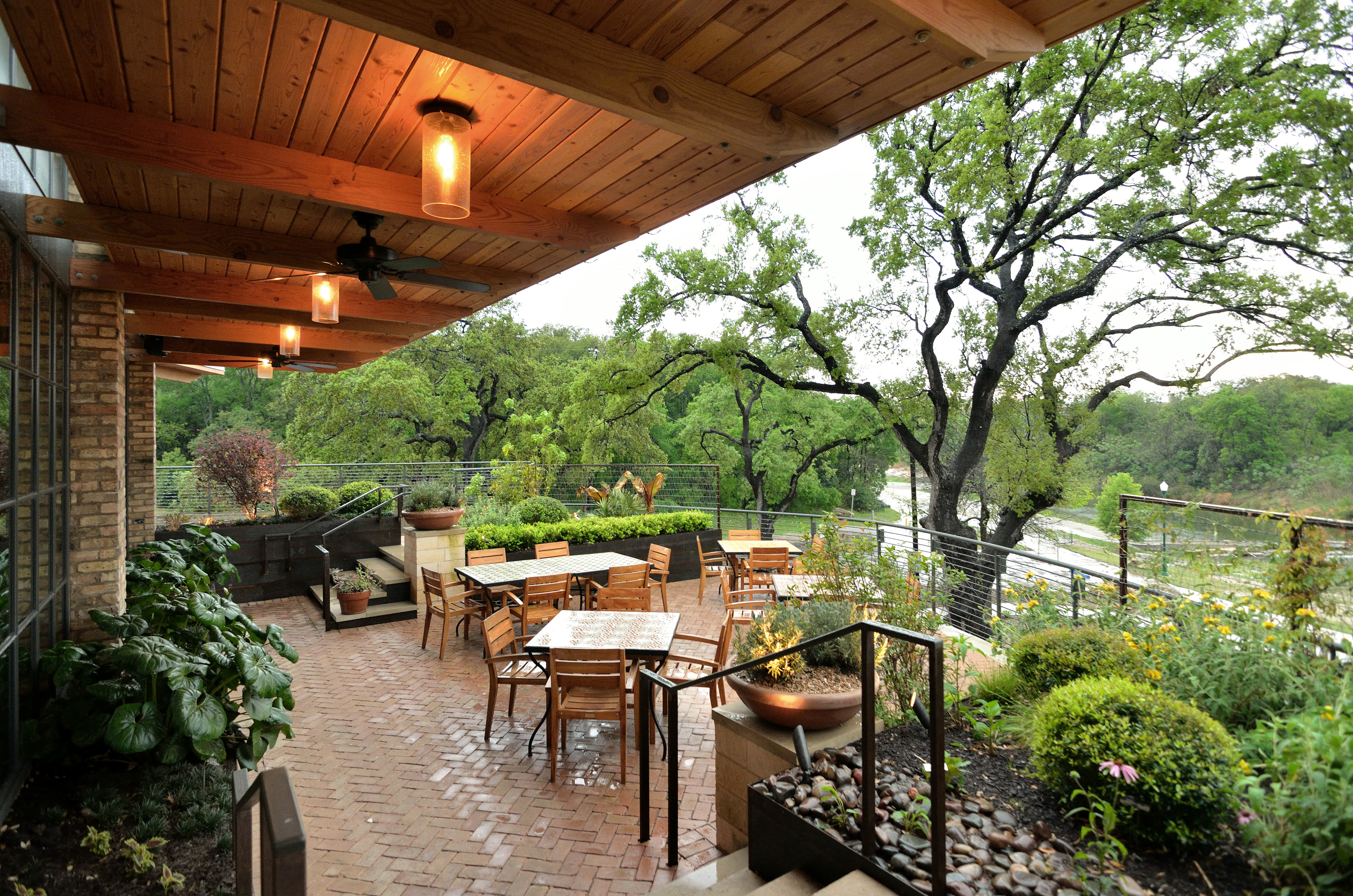 A covered outdoor patio with wooden tables and chairs, surrounded by greenery, features overhead lights and a view of a wooded area in the background, making it an ideal spot for gatherings that feel refreshingly disconnected from the usual auto draft of daily life.