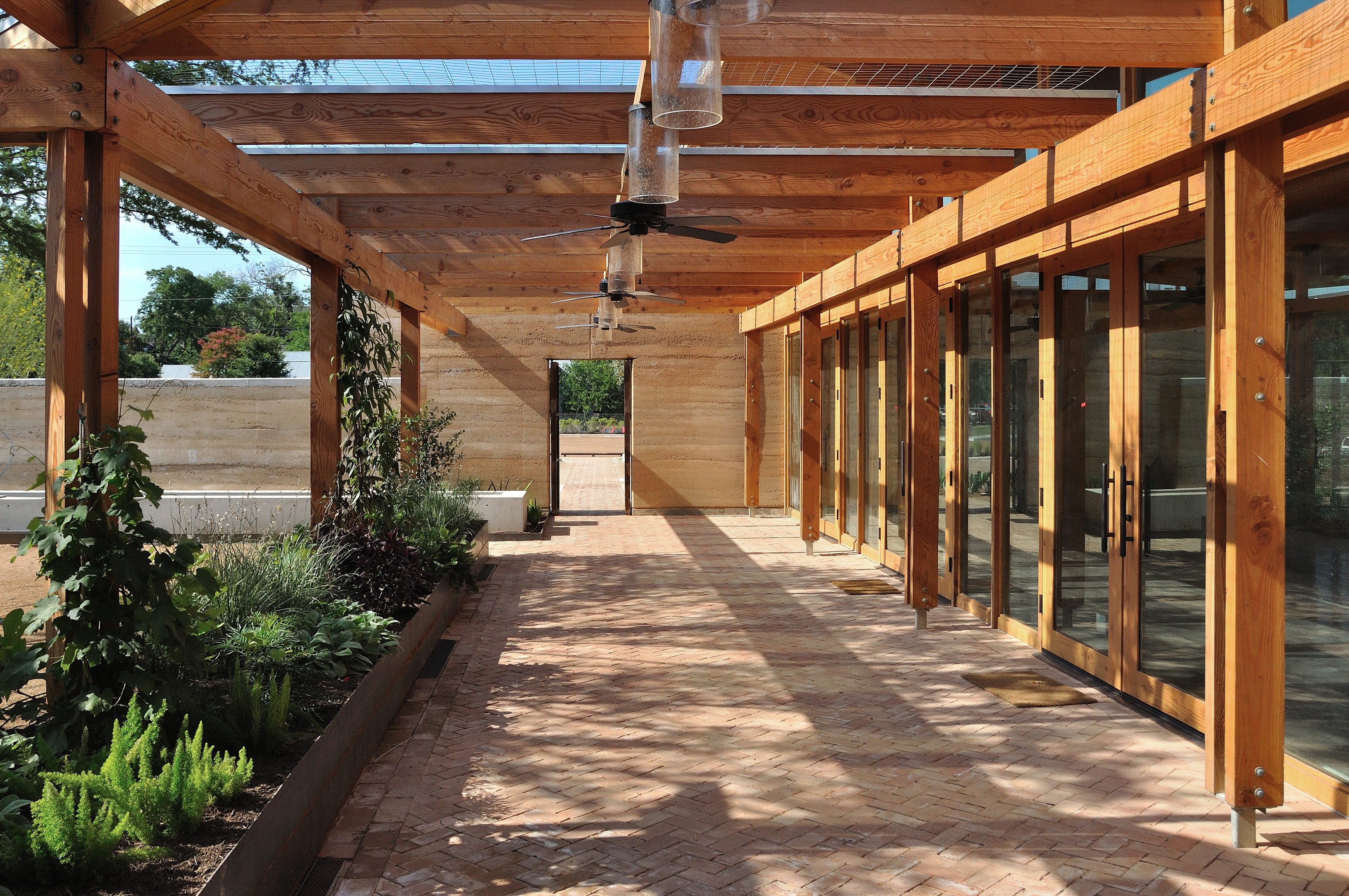 A spacious, sunlit terrace with wooden beams, brick flooring, glass doors, ceiling fans, and an entranceway. Green plants line the left side while trees are visible in the background. Whether you’re relaxing or working on an Auto Draft project, this terrace offers a serene space for both.