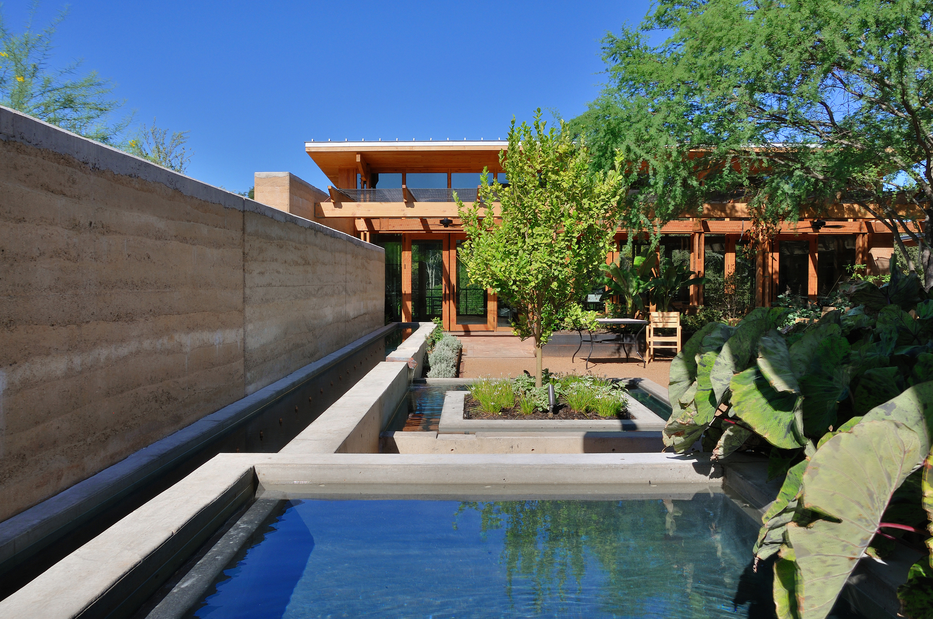 Modern house with large windows, surrounded by greenery and featuring a reflective water feature in the foreground. The clear blue sky adds a touch of serenity, seamlessly blending the natural and designed elements as if an auto draft of tranquility.