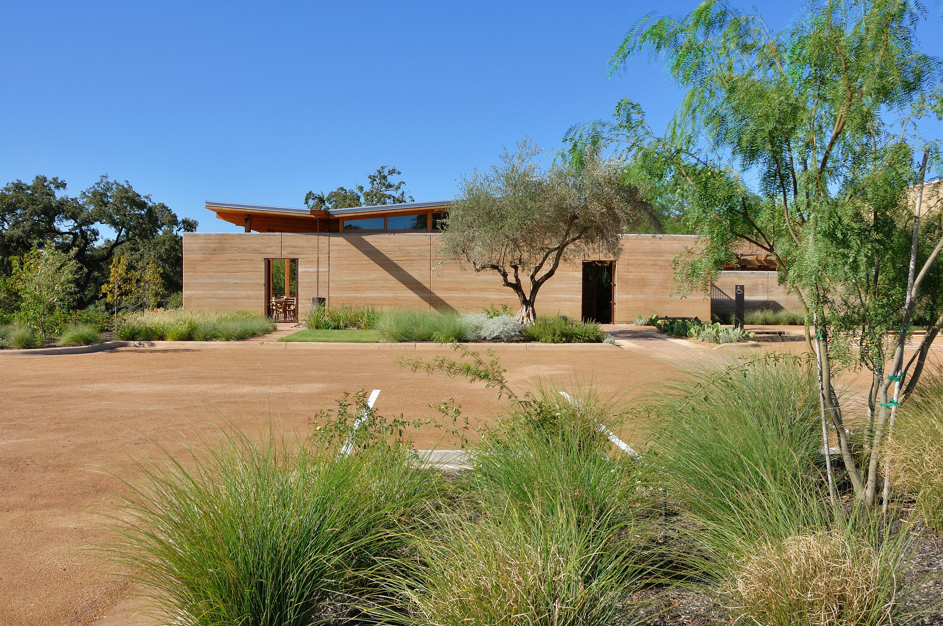 A single-story modern building with large windows and a flat roof is surrounded by landscaped greenery and an auto draft dirt parking area.