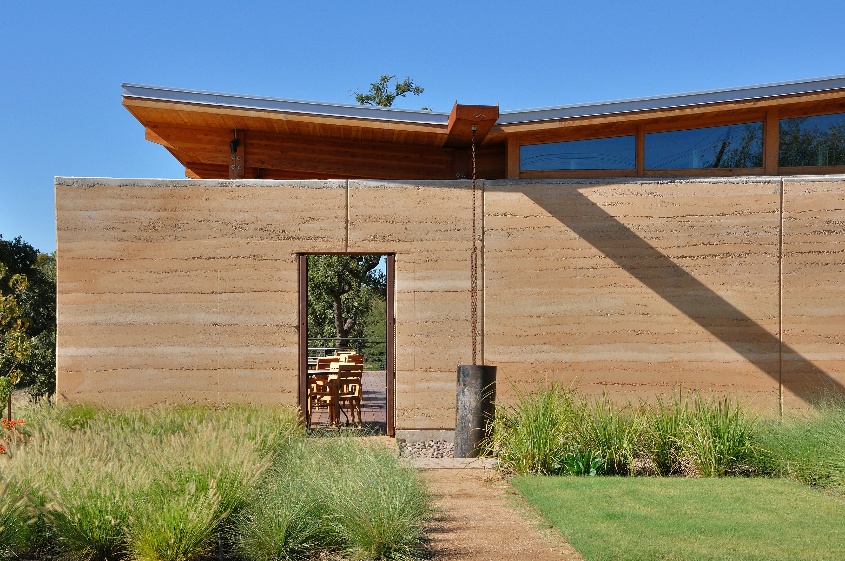 A modern building with a flat roof and large windows, featuring a rustic earthen wall. An open doorway leads to an outdoor seating area, surrounded by lush greenery—an auto draft of architectural brilliance.