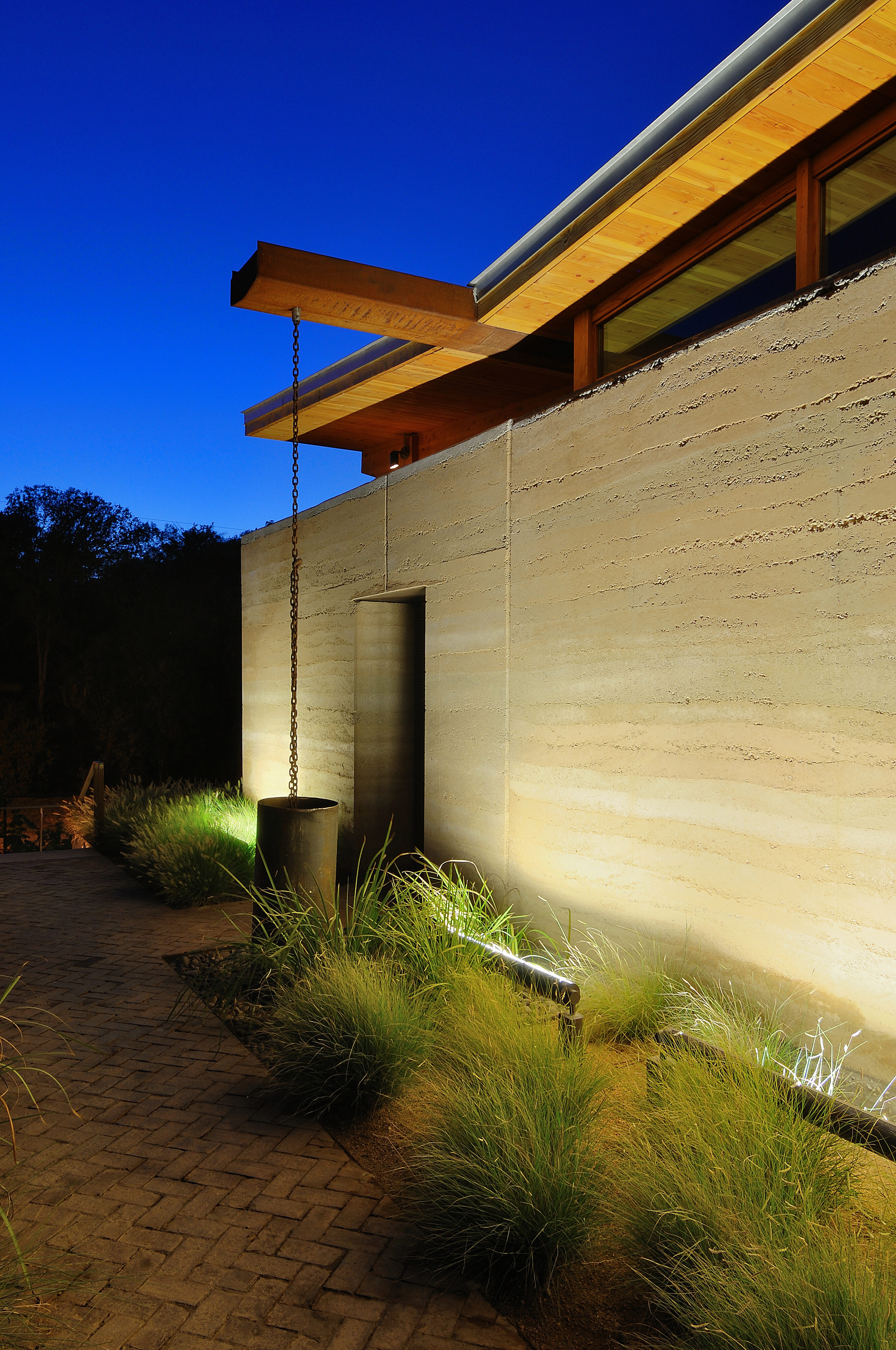 A modern building exterior at dusk showcases an auto draft chain rain gutter, an illuminated stone wall, and plants lining the paved walkway.