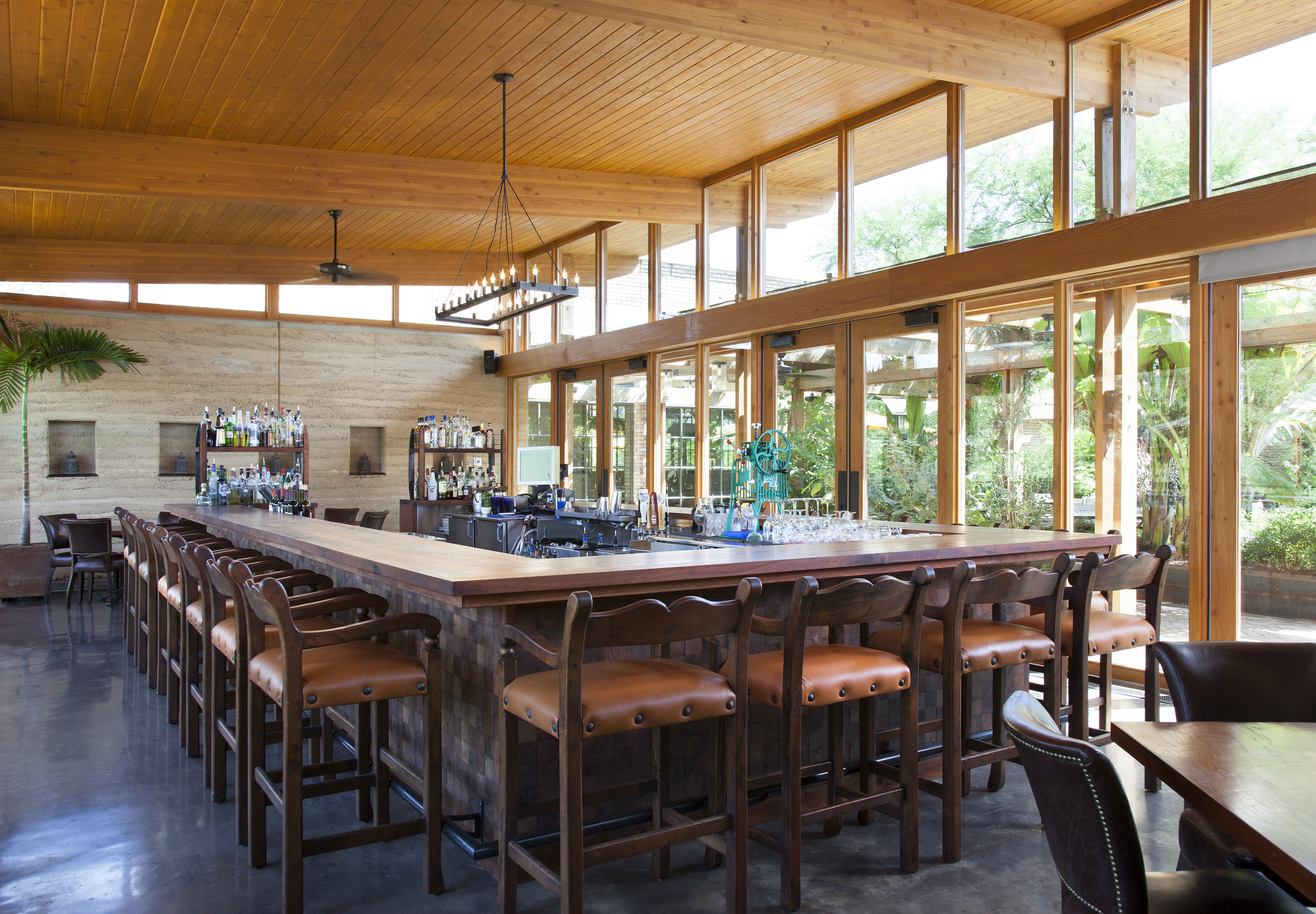 Spacious bar area with wooden stools around a large central counter. The room, akin to an auto draft of rustic charm, features large windows, a wooden ceiling, and various bottles displayed on shelves behind the bar.