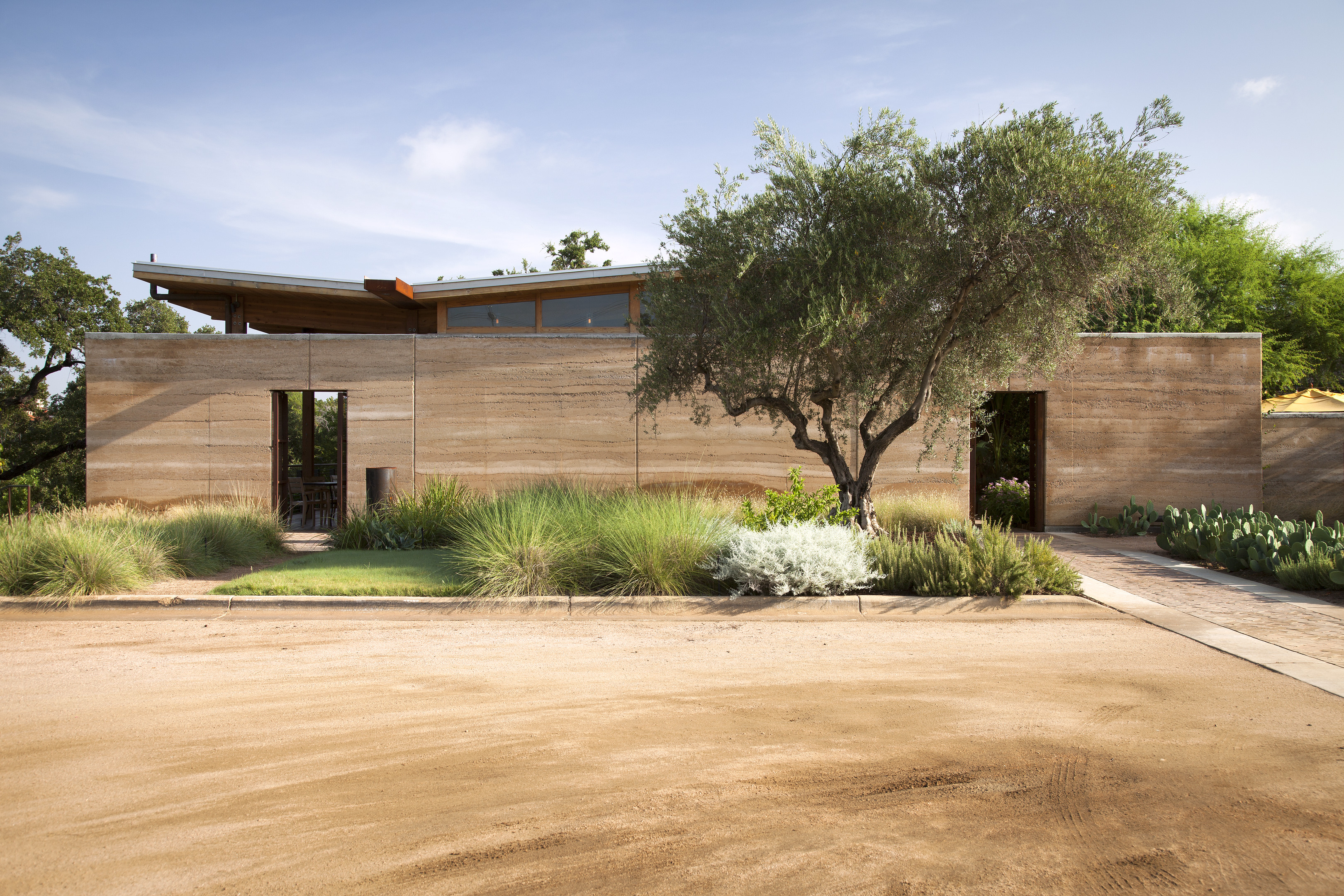 A modern house with a flat, rectangular facade made of brown earth-toned materials features a meticulous auto draft design. In front, there is a dirt path, greenery, and a tree. The sky is clear.