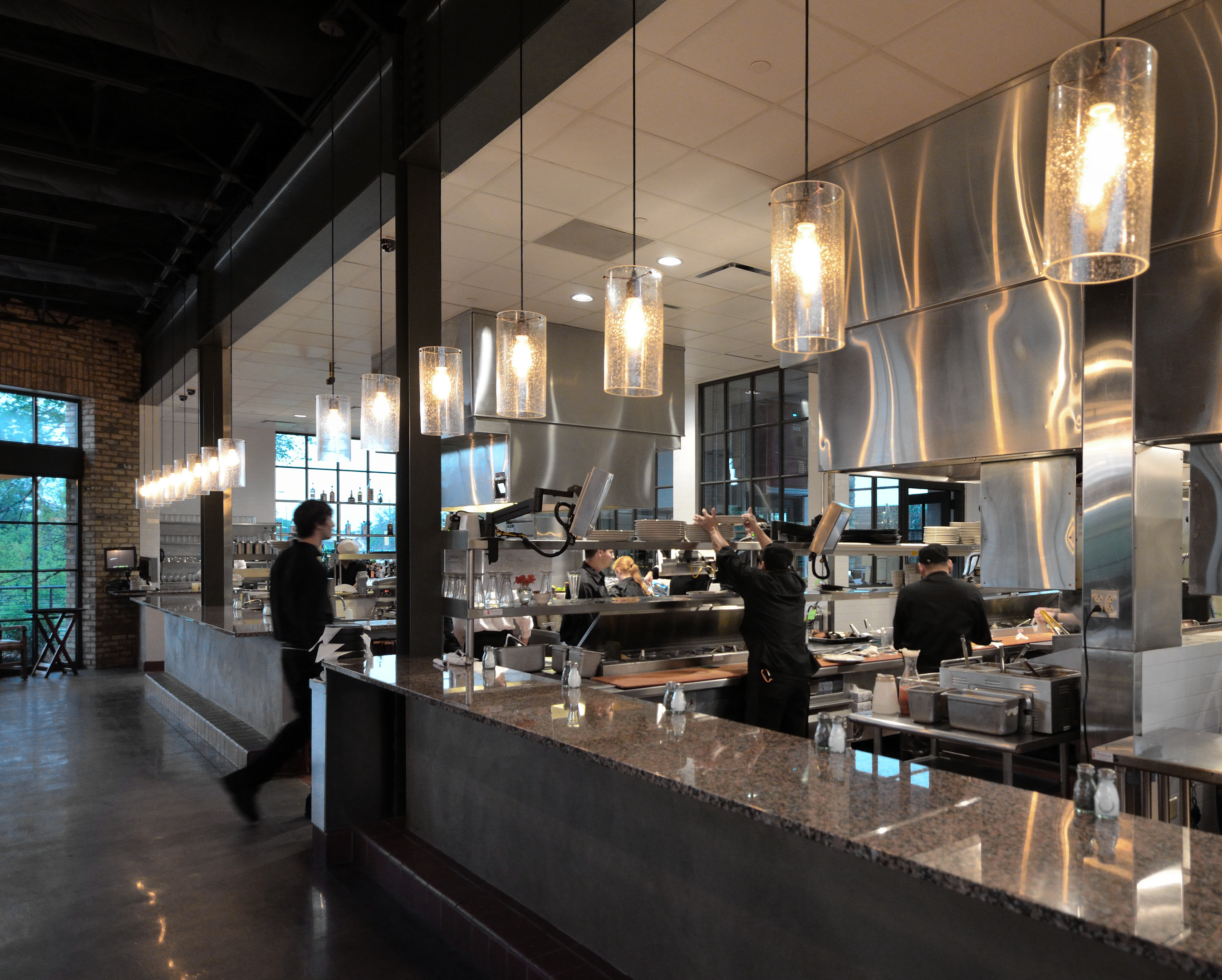 A modern, open kitchen in a restaurant with chefs preparing food and a staff member walking by. Hanging pendant lights illuminate the space, creating an atmosphere so well-orchestrated it feels like an auto draft of culinary excellence.