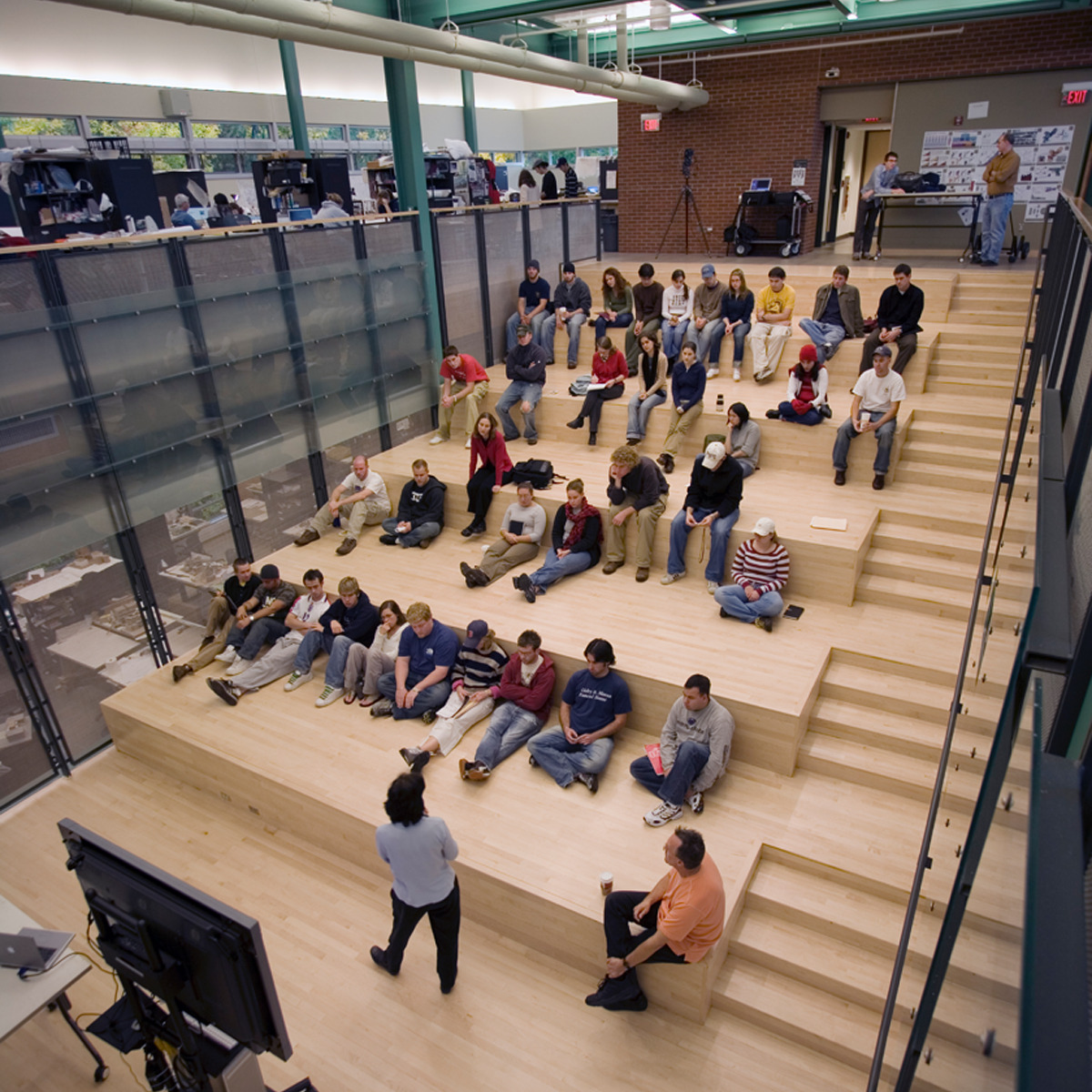 A group of people seated on tiered wooden steps attentively listen to a speaker at the bottom. The setting appears to be a modern, industrial-style conference or educational space, possibly within the University of Texas’ Student Activity Center.