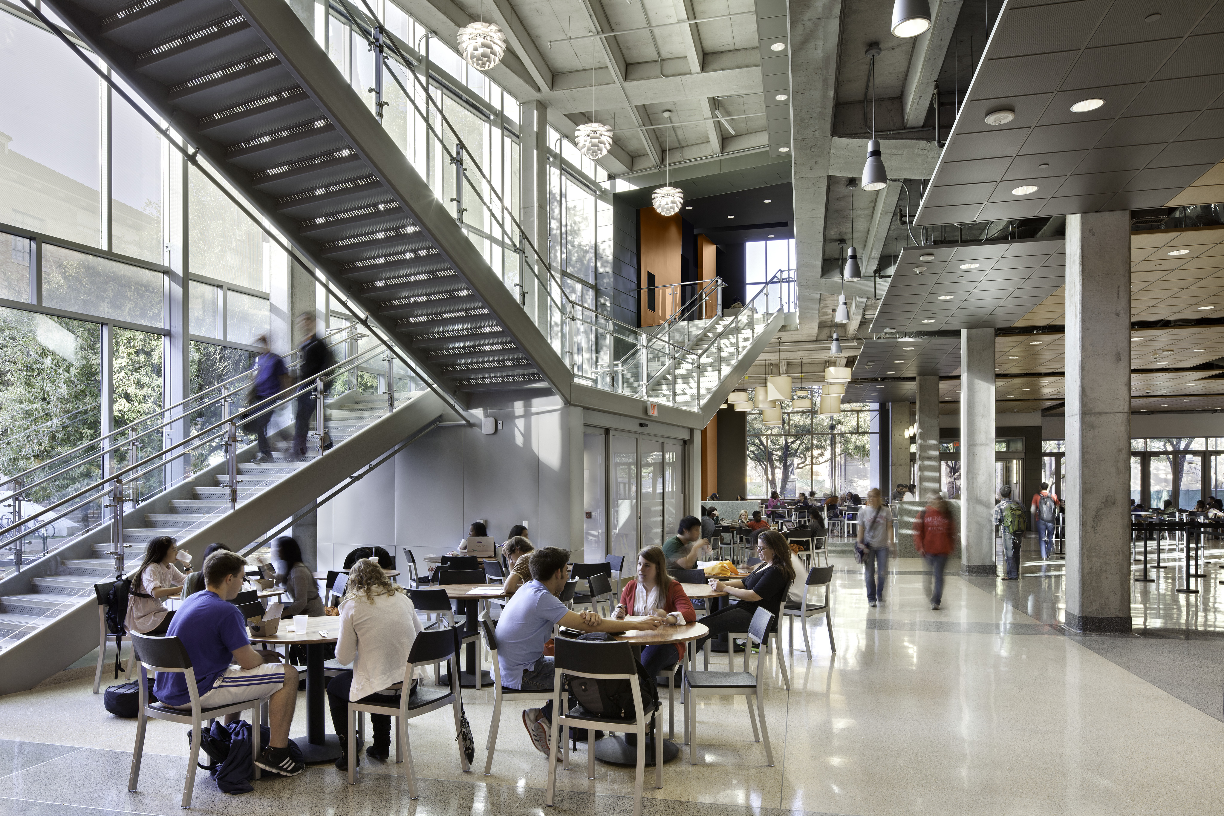 A modern cafeteria in the University of Texas Student Activity Center features large windows, high ceilings, and people sitting at tables. A staircase leads to an upper level, and several individuals are walking around the space.