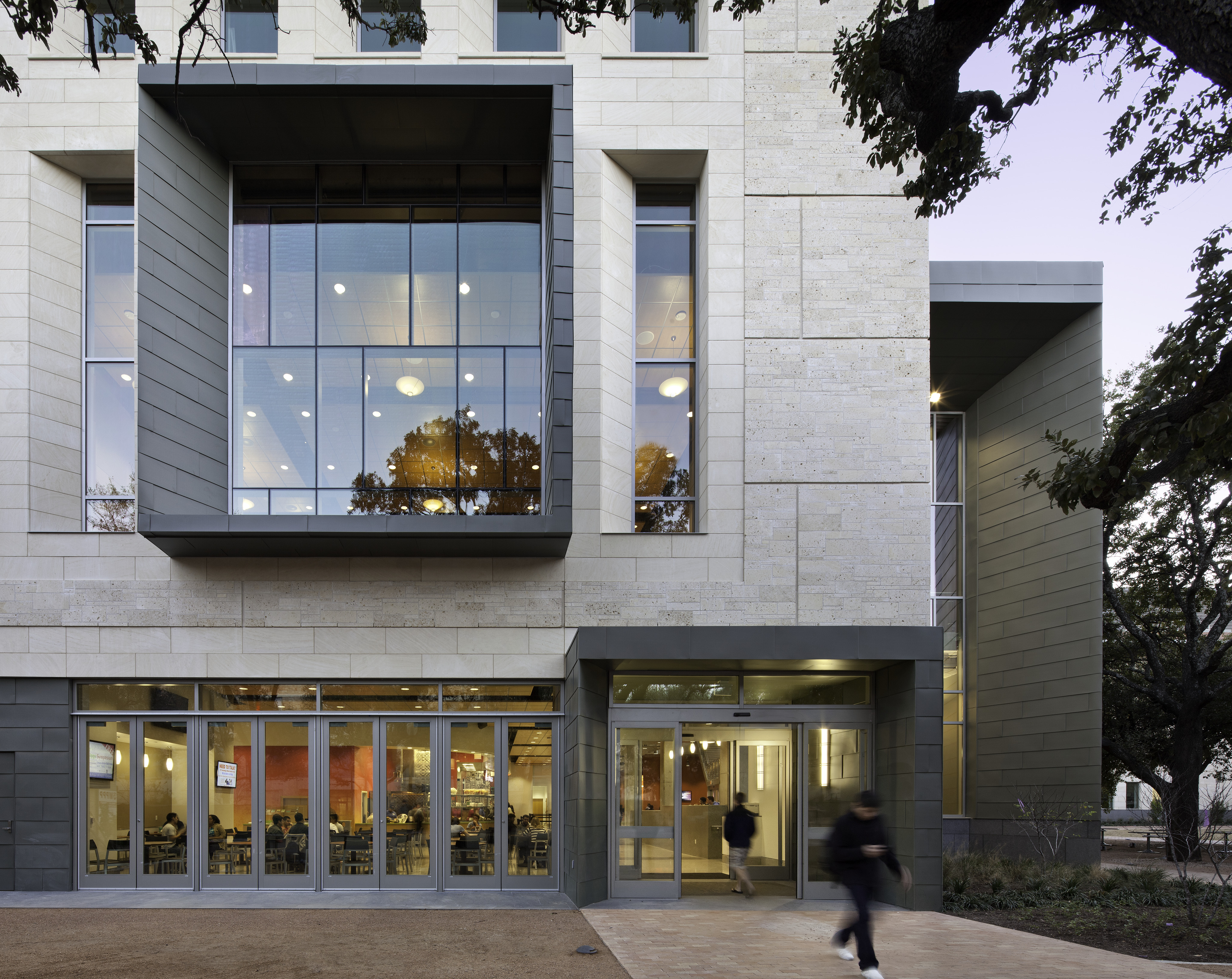 Modern building facade with large windows at the University of Texas. One person walking into the entrance, another blurred in motion just outside. Trees and dusk sky visible in reflections and background, marking a vibrant evening at the Student Activity Center.