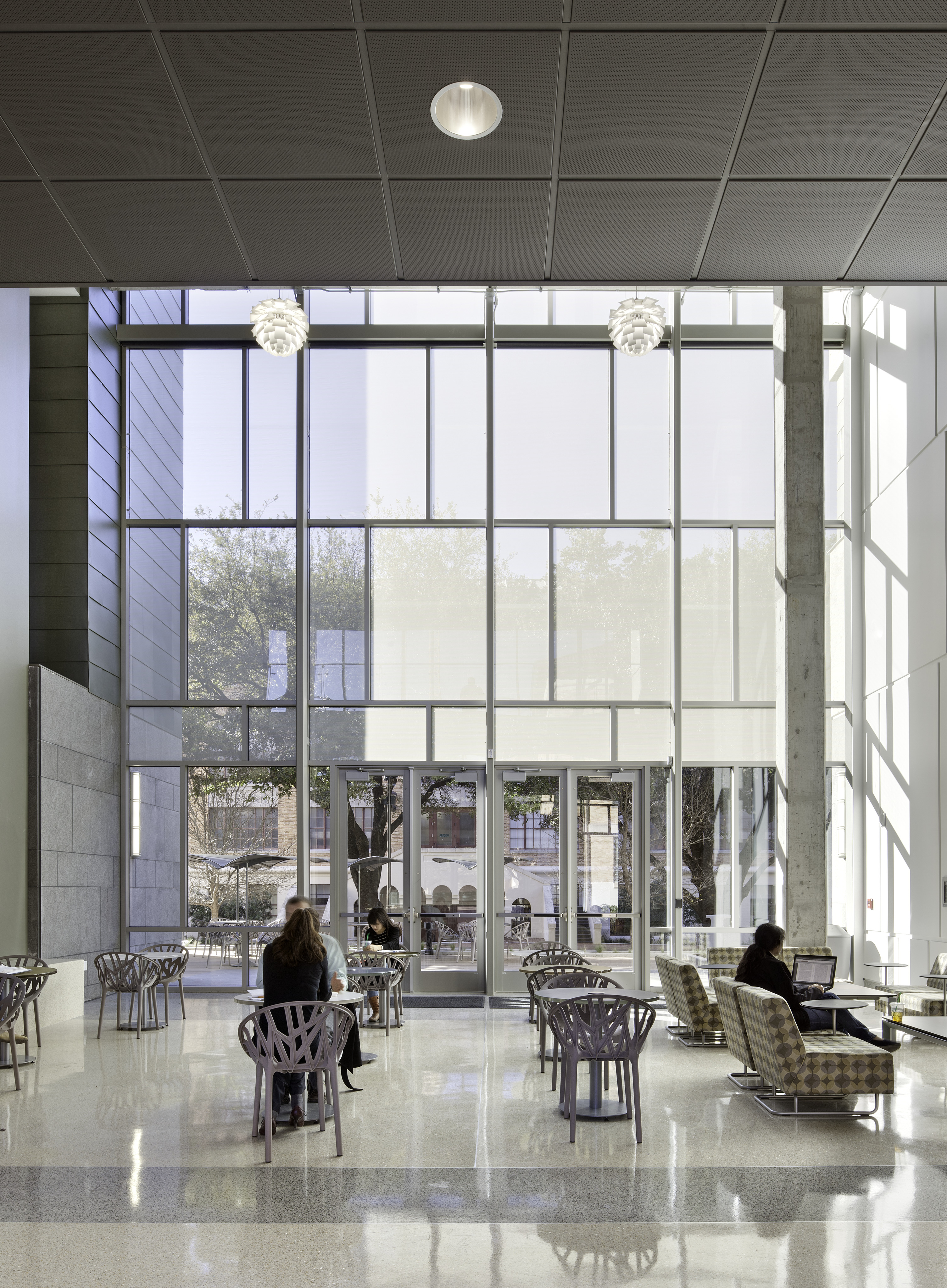 A modern, well-lit interior space with high ceilings and large windows at the University of Texas. Several people are seated at tables and in chairs, using laptops and engaging in conversation, creating a vibrant atmosphere typical of a Student Activity Center.