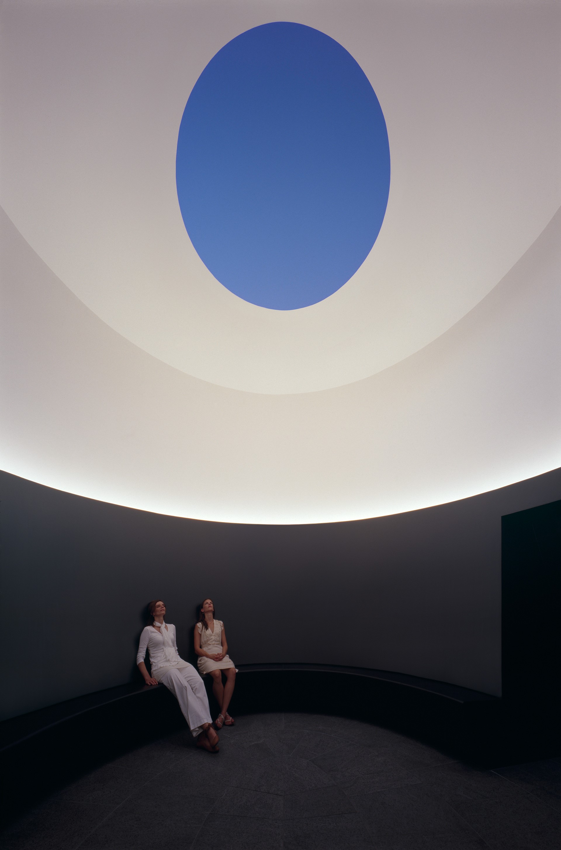 Two people sit on a curved bench in a room with a circular skylight revealing a blue sky, experiencing an immersive art installation reminiscent of a James Turrell Skyspace.