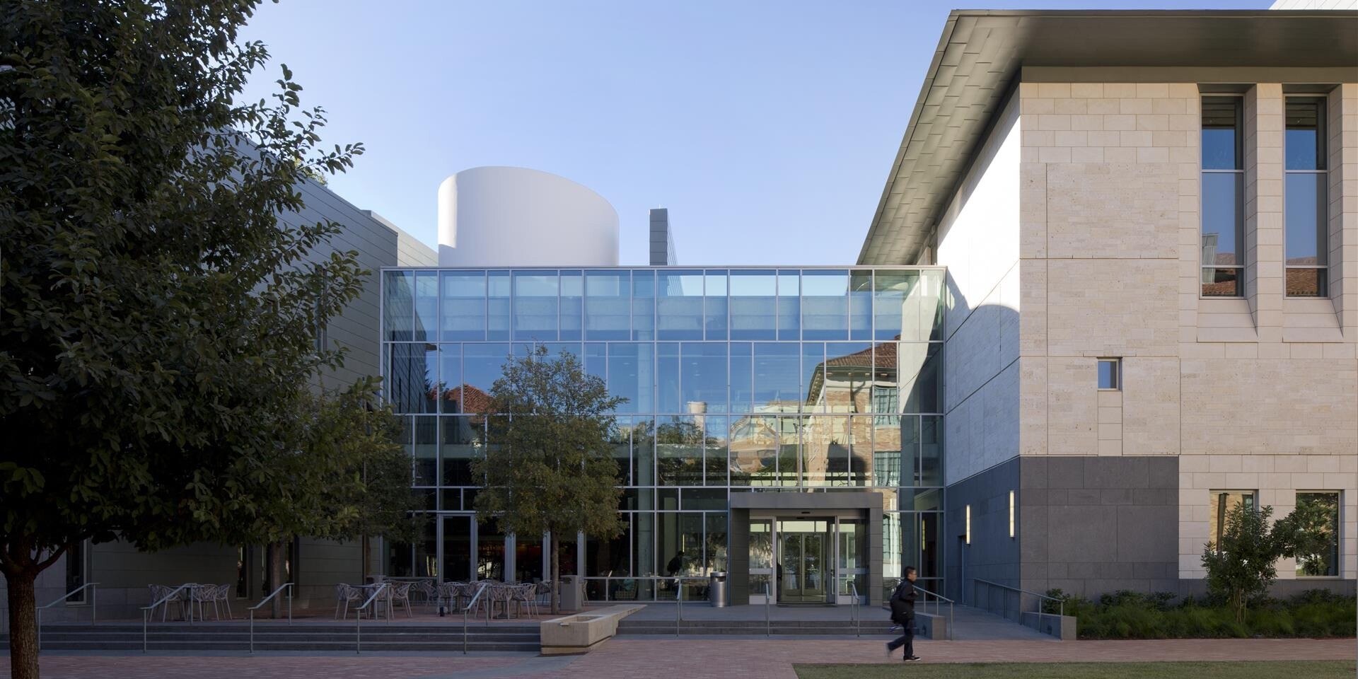 Modern Student Activity Center at the University of Texas with a glass façade, outdoor seating area, and landscaped greenery in front. Person walking on the pathway under a clear blue sky.