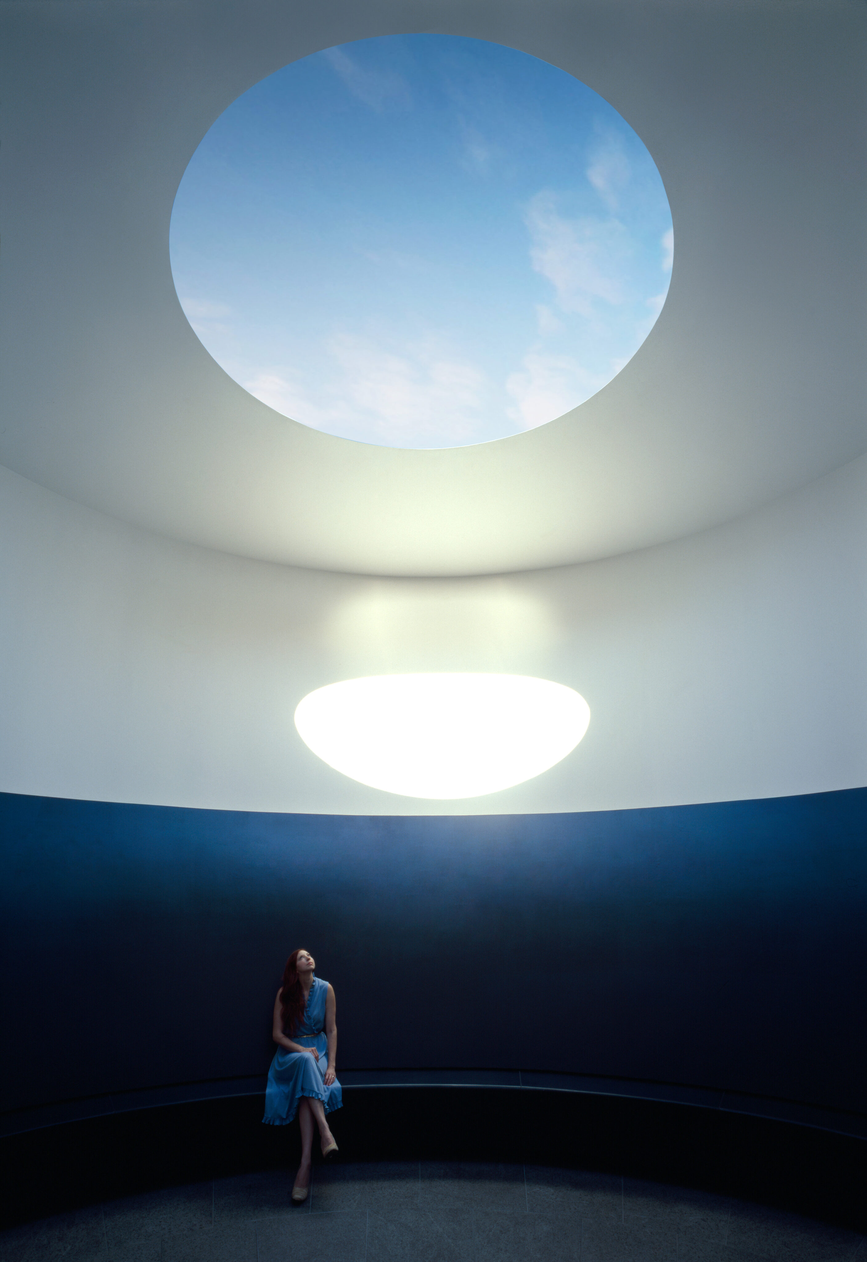 A woman in a blue dress sits on a curved bench in a minimalist room, reminiscent of a James Turrell Skyspace, with a large circular skylight illuminating the space and adding color inside.