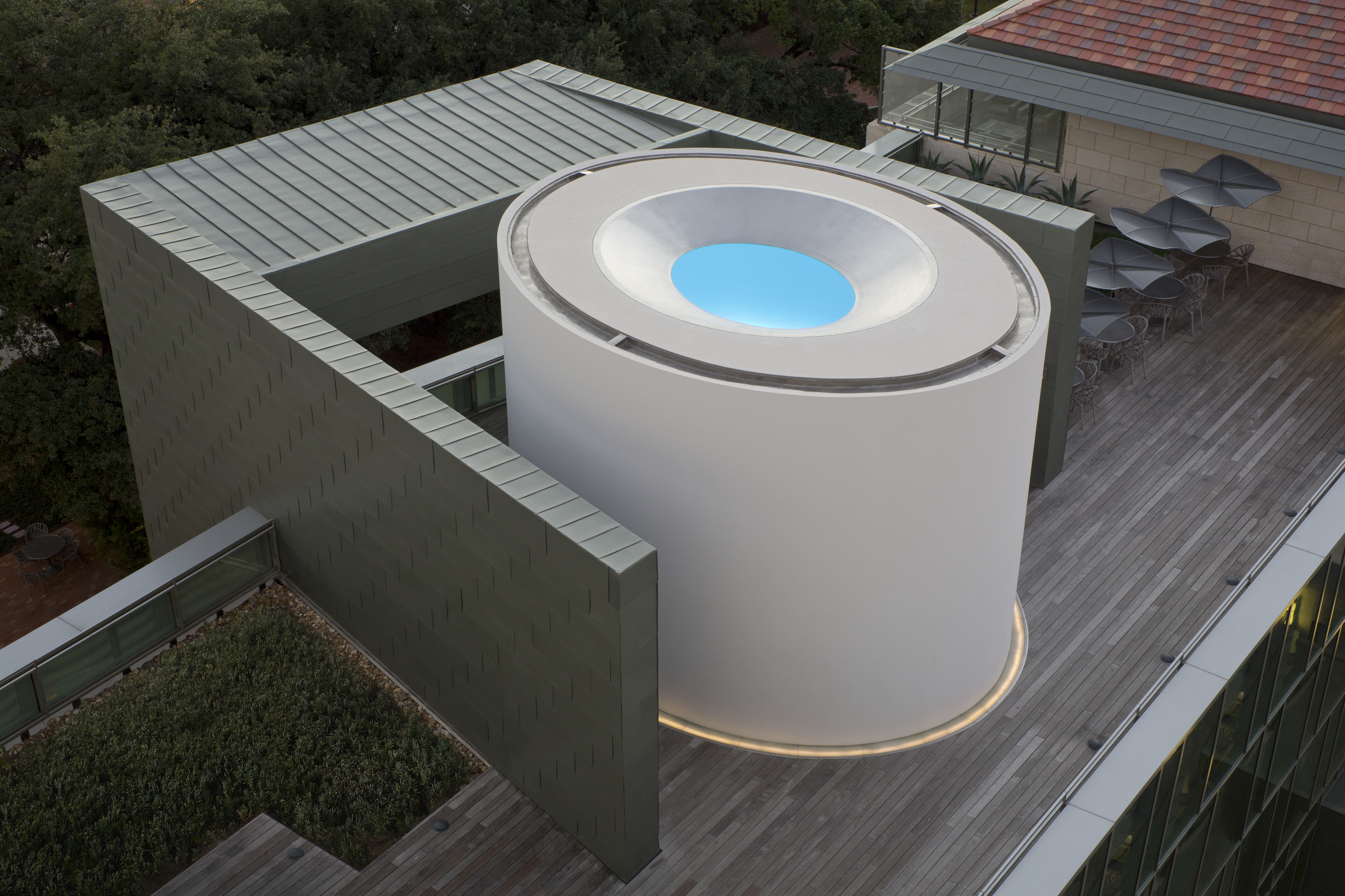 A cylindrical building with an open roof pool, reminiscent of James Turrell's Skyspace installations, is positioned on a rooftop deck surrounded by green walls and adjacent structures, with outdoor seating visible nearby.