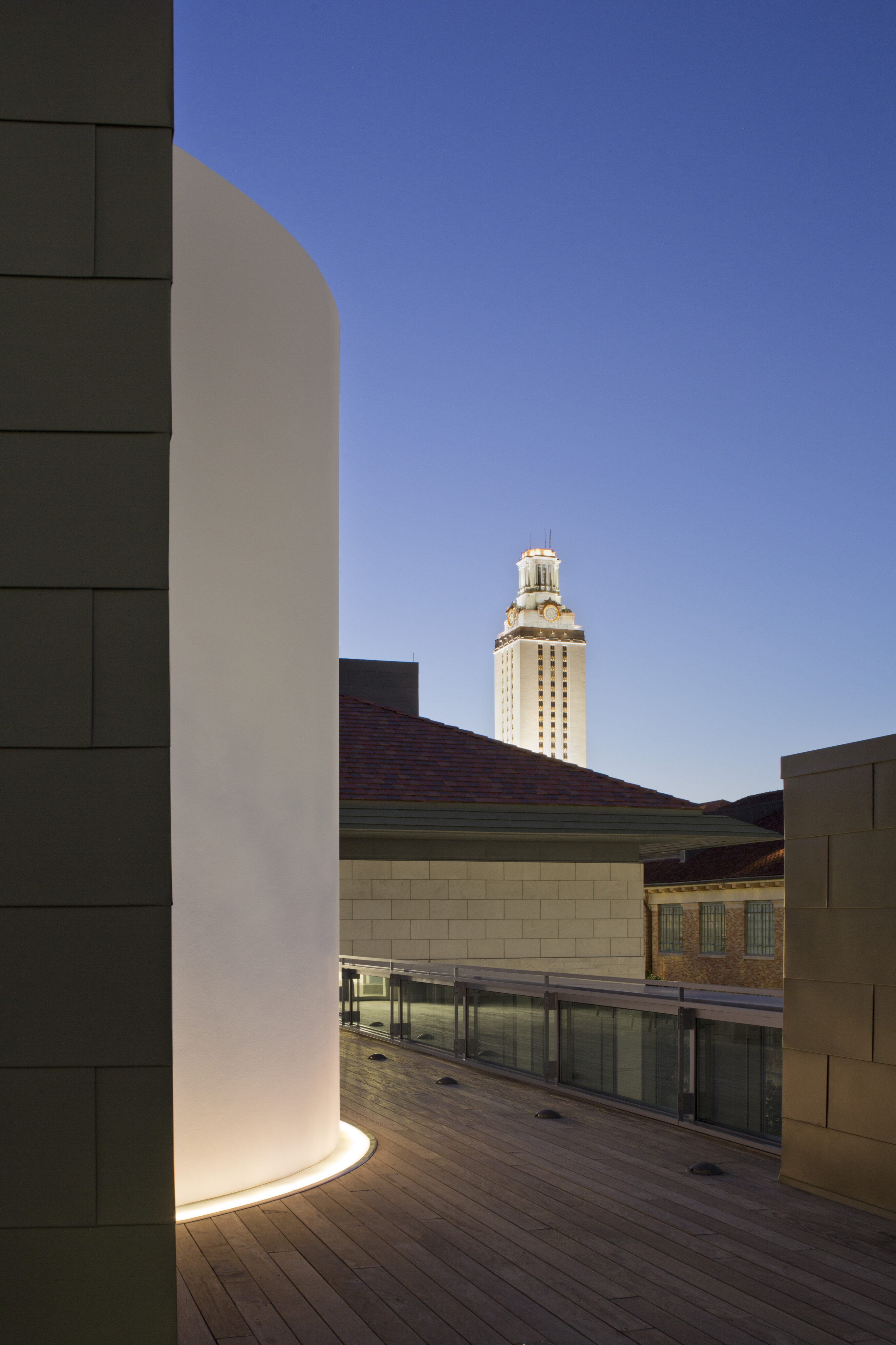 A tall clock tower is visible in the distance against a clear evening sky, seen from a rooftop with modern architectural features and a wooden deck walkway illuminated by ground-level lights, reminiscent of a James Turrell Skyspace.