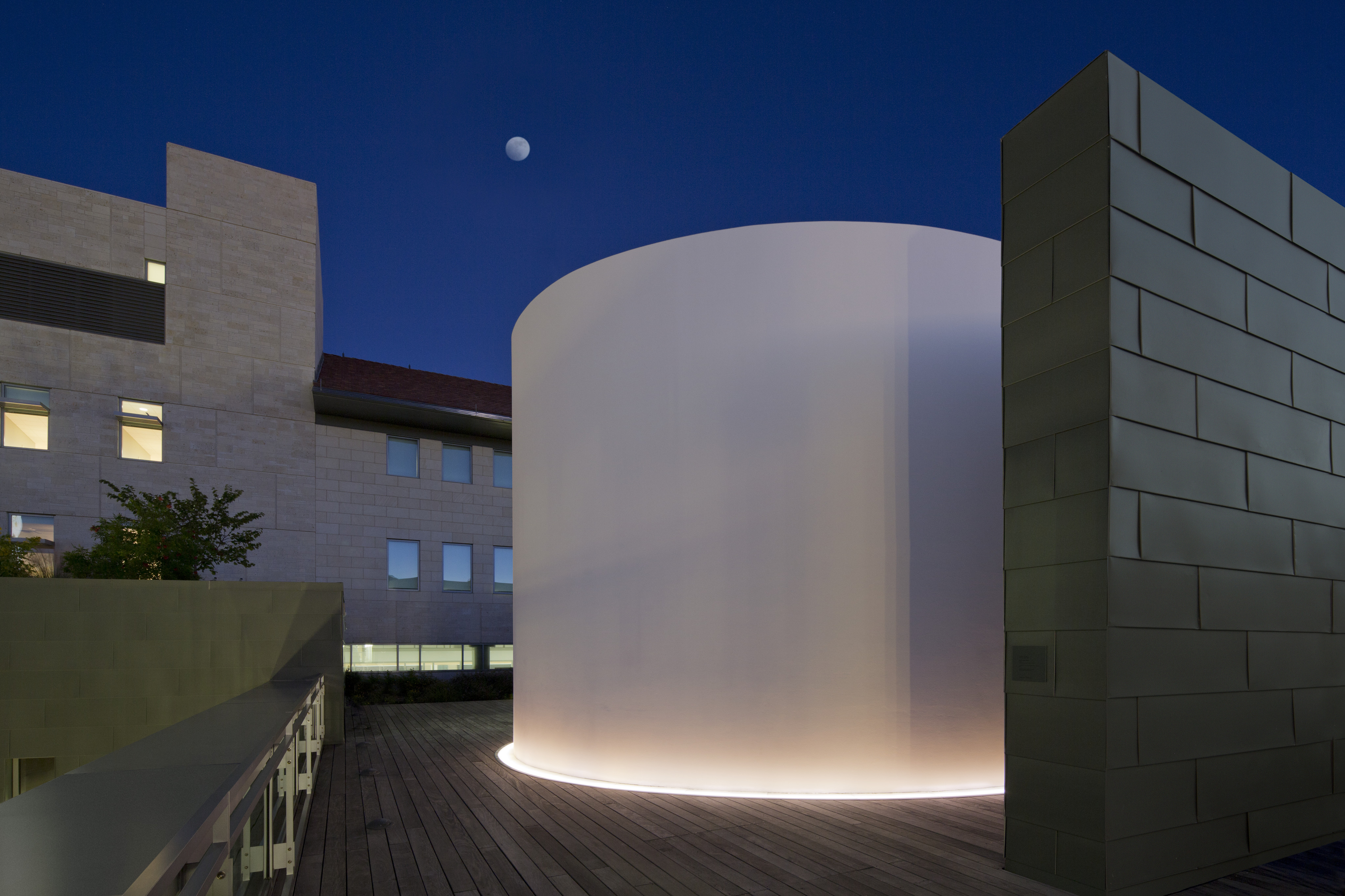 Modern architectural structure with illuminated cylindrical feature at night, accompanied by nearby buildings and a visible moon in the dark sky, reminiscent of James Turrell’s Skyspace installations like The Color Inside.