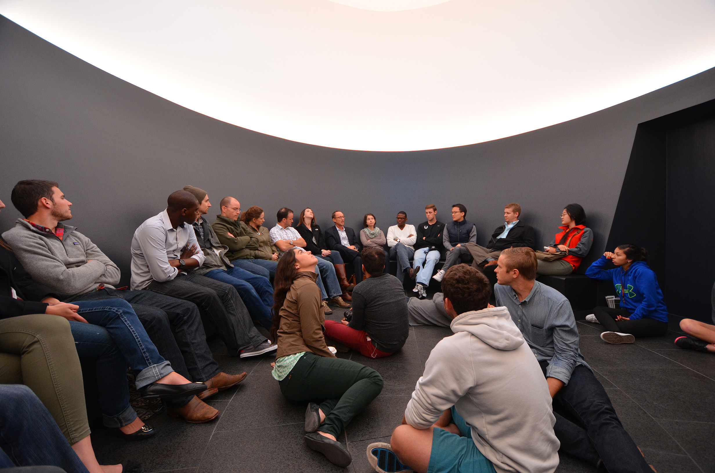 A group of people sits in a semi-circle in a modern, minimalist room with curved walls and indirect lighting. Some participants sit on chairs while others are seated on the floor, reminiscent of a James Turrell Skyspace art installation titled "Color Inside.