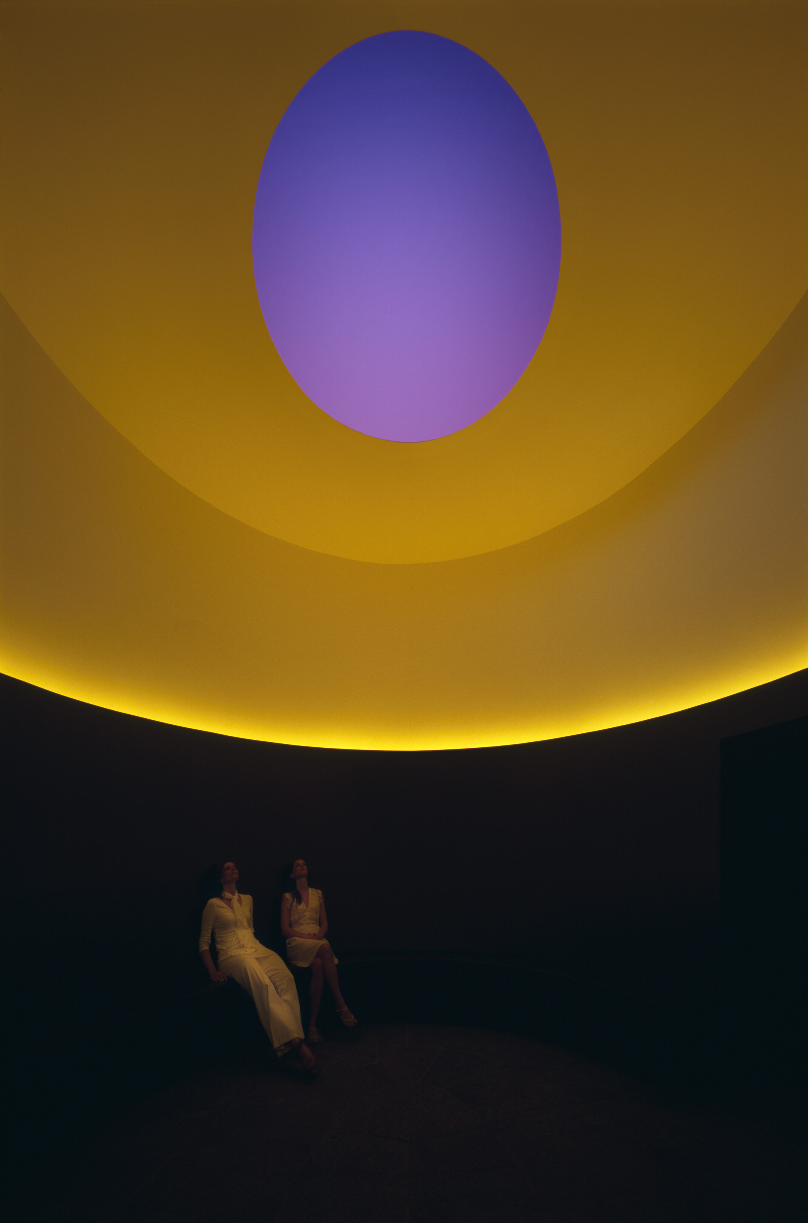 Two people sit in a dark room illuminated by yellow and purple light emanating from a circular Skyspace structure in the ceiling.