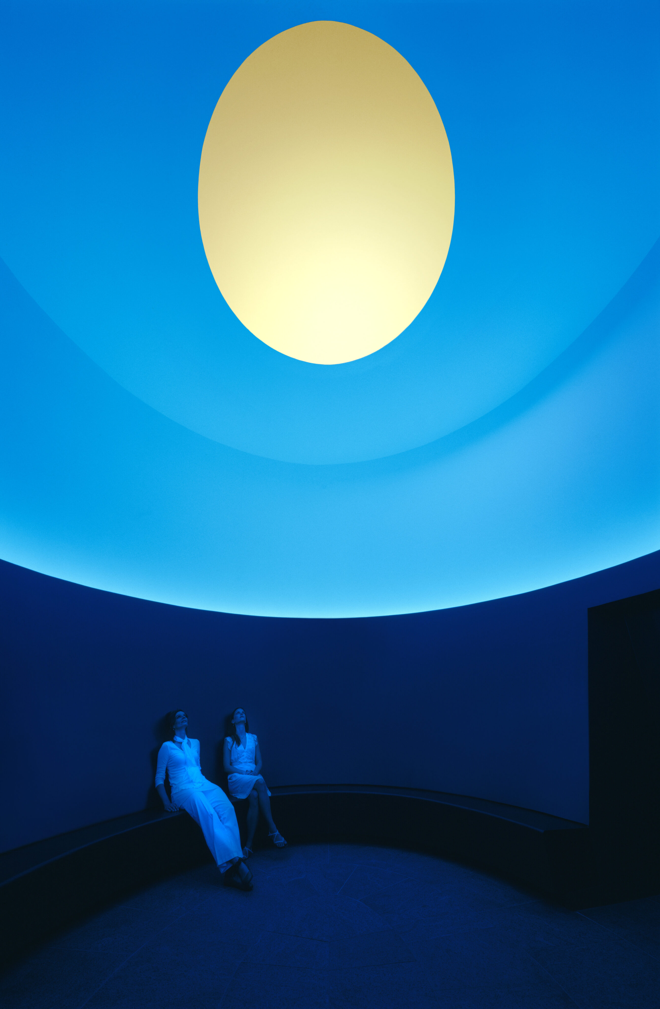 Two individuals in white attire sit on a curved bench in a room with blue walls, beneath a large circular light resembling the sun, reminiscent of James Turrell's Skyspace installations like The Color Inside.