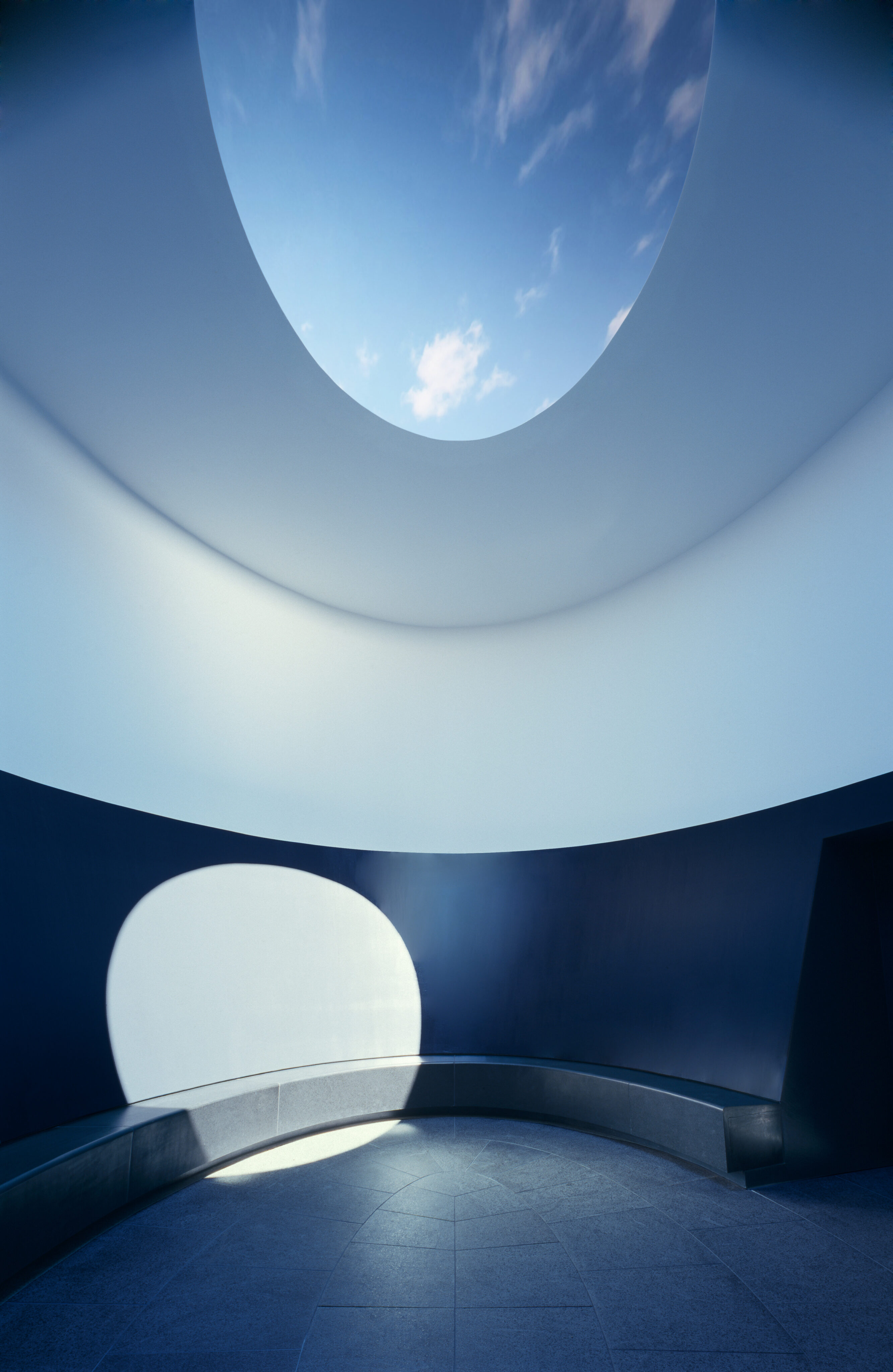 A minimalistic circular room with curved walls, an open ceiling revealing the sky, and a bench along the wall, reminiscent of a James Turrell Skyspace.