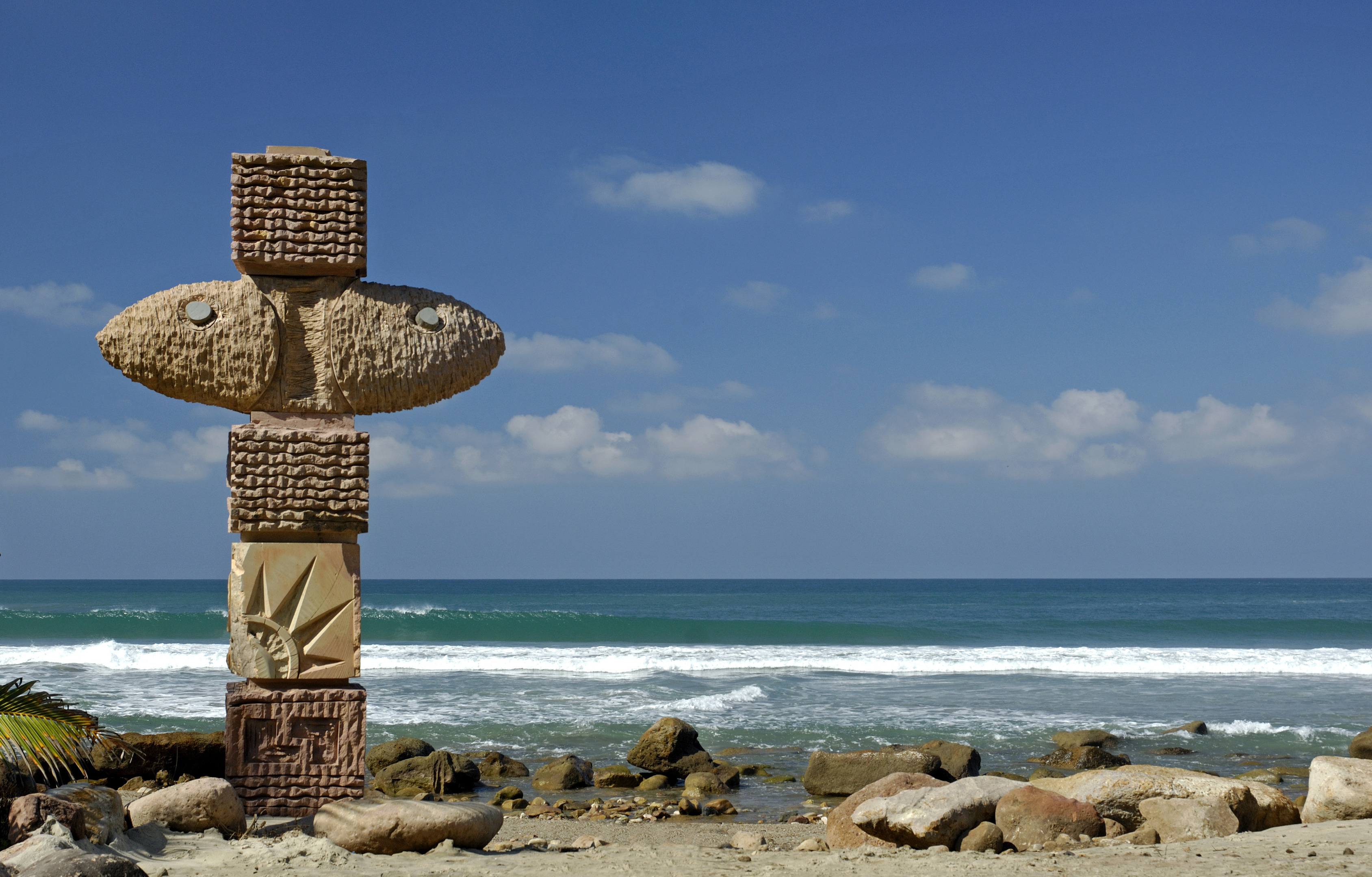 A totem pole stands on the rocky beach of Imanta Resort, with ocean waves and a clear blue sky in the background.