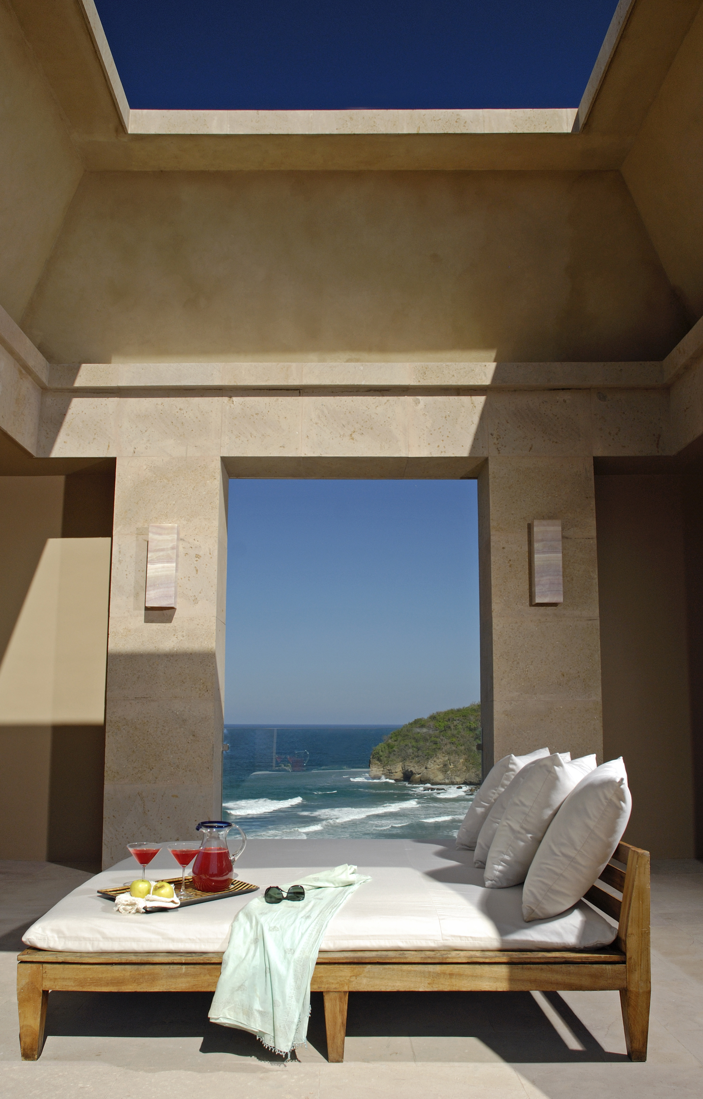 A lounge chair with a green towel, sunglasses, drinks, and snacks awaits you at Imanta Resort, right in front of an open doorway with a breathtaking view of the ocean and sky.