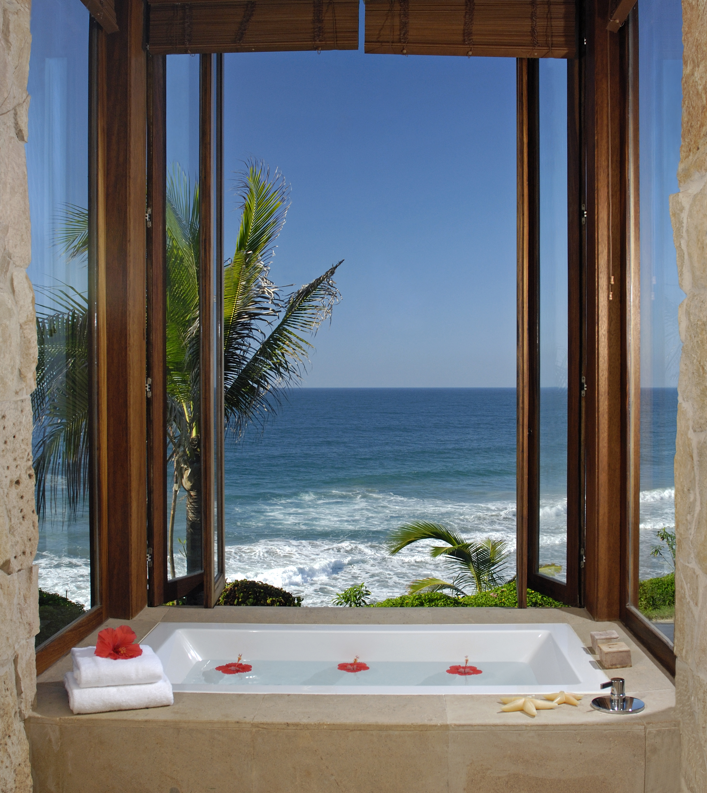 A bathtub with floating flowers is positioned near open windows overlooking the ocean and palm trees at Imanta Resort. White towels and a starfish adorn the edge of the tub, creating a serene spa-like retreat.