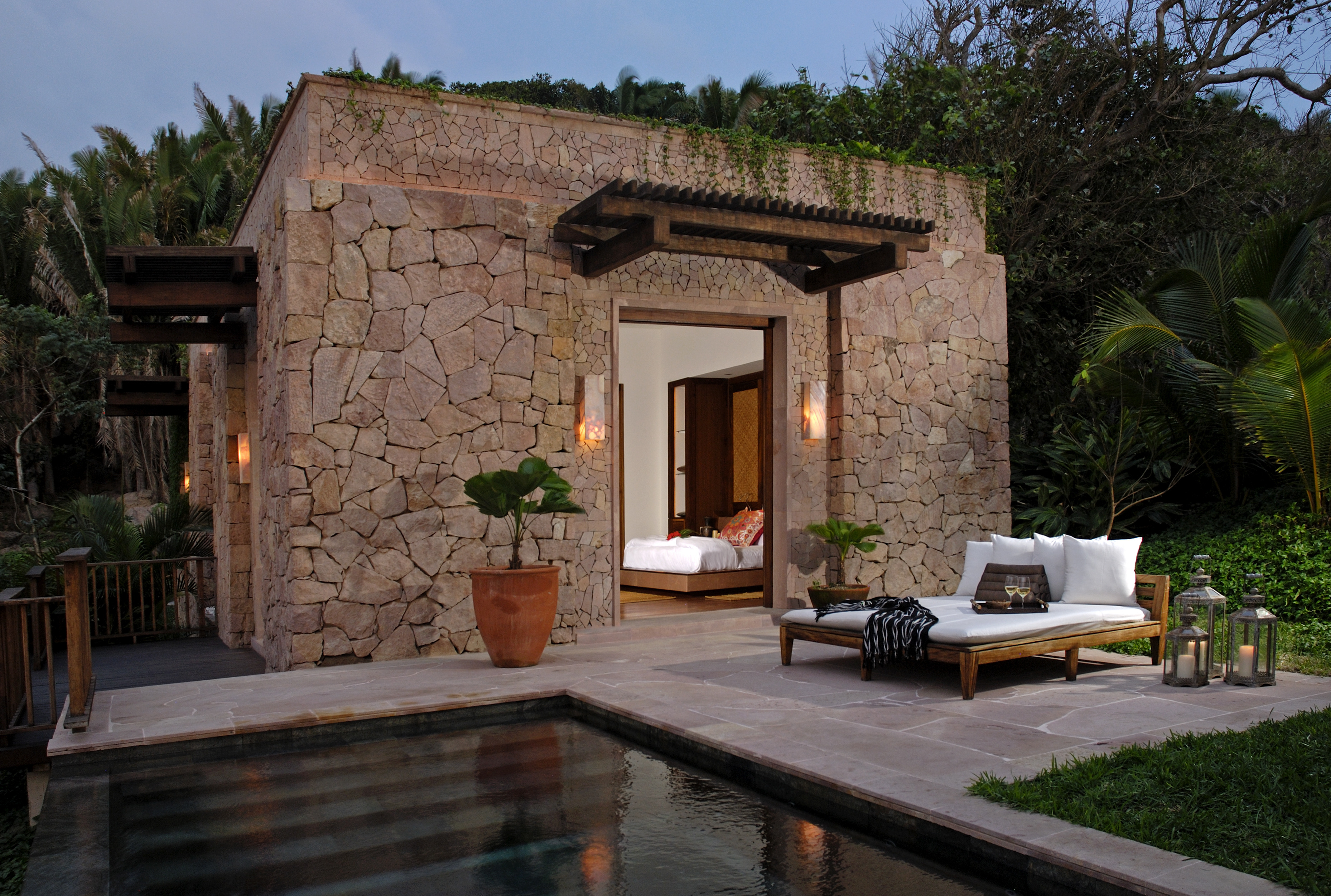 A stone cottage with a pergola-covered entrance, seen at dusk with a glimpse of a bedroom inside, evokes the tranquility of an Imanta resort. A small pool, lounge chairs, and tropical plants enhance this spa-like retreat.