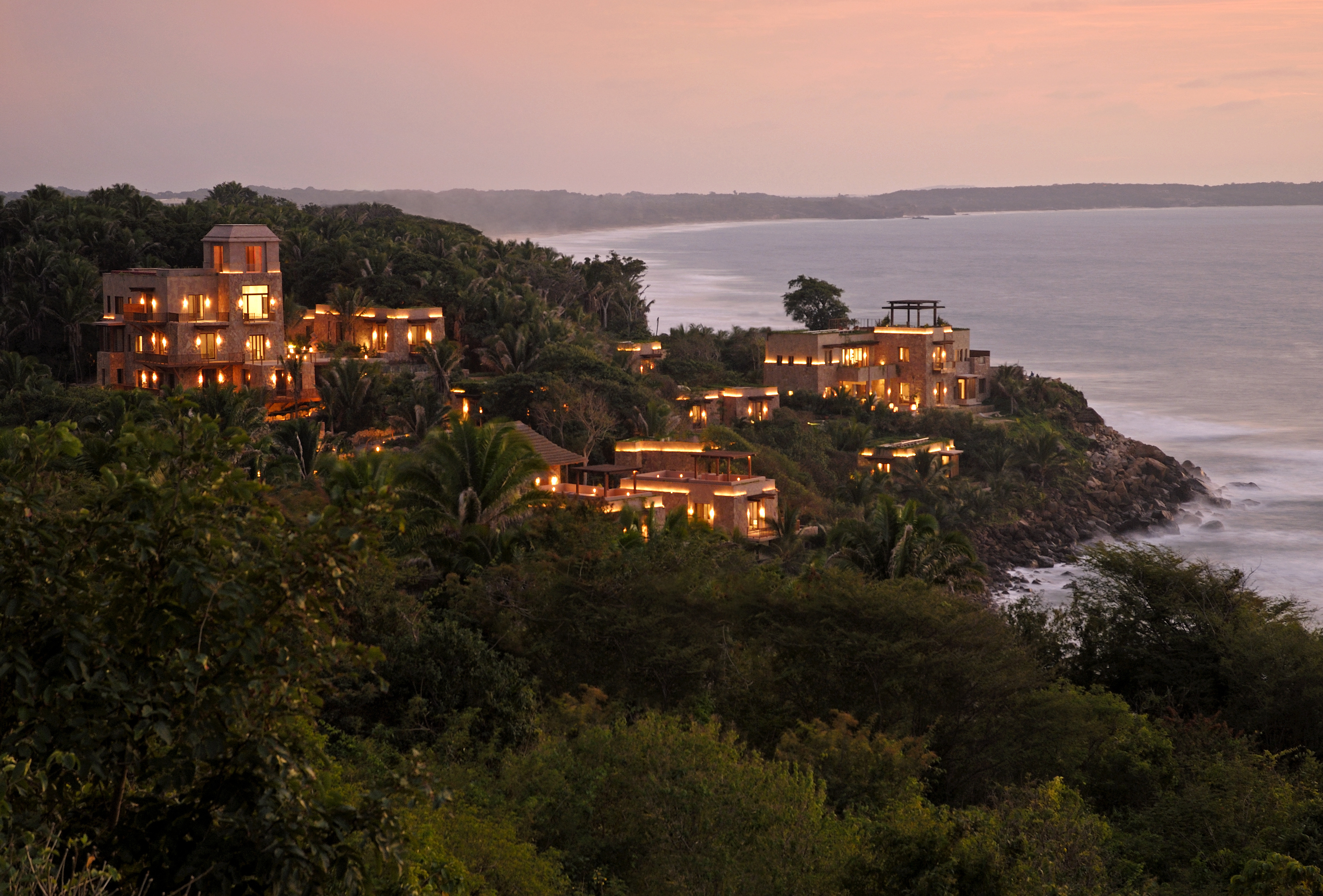 A coastal resort at sunset, featuring multiple lit-up buildings nestled among lush greenery, overlooking the ocean—welcome to Imanta Resort and Spa.