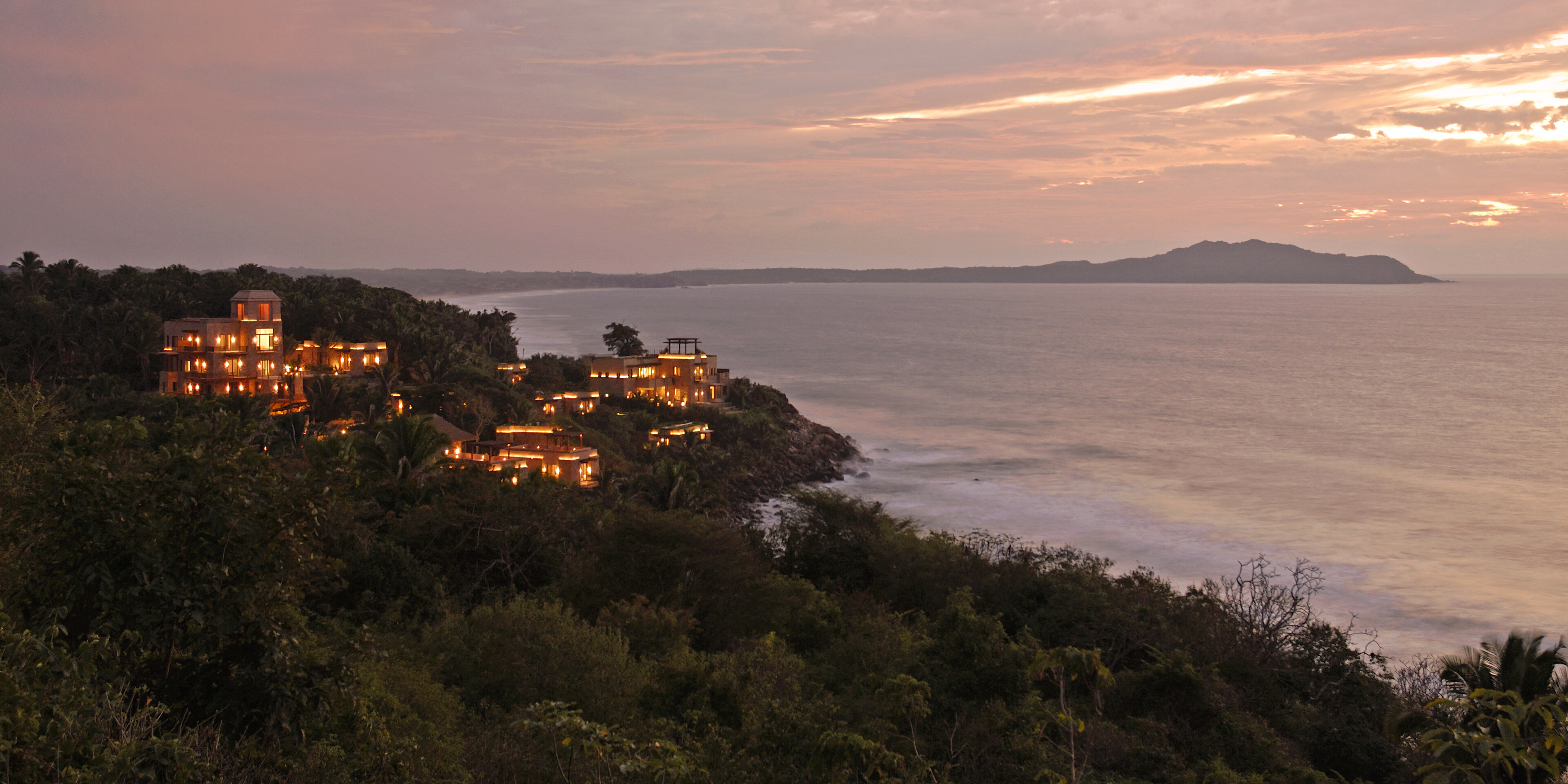 Sunset view of a coastline with buildings nestled among the hills, their lights glowing. An expansive ocean and an island in the distance are visible under a partly clouded sky, perfect for those seeking tranquility at Imanta Resort.