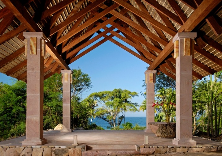 Open-air wooden pavilion with a triangular roof, supported by stone pillars, overlooks lush greenery and the ocean in the background, making it an ideal spot within Imanta Resort to enjoy the serene surroundings or unwind at the spa.