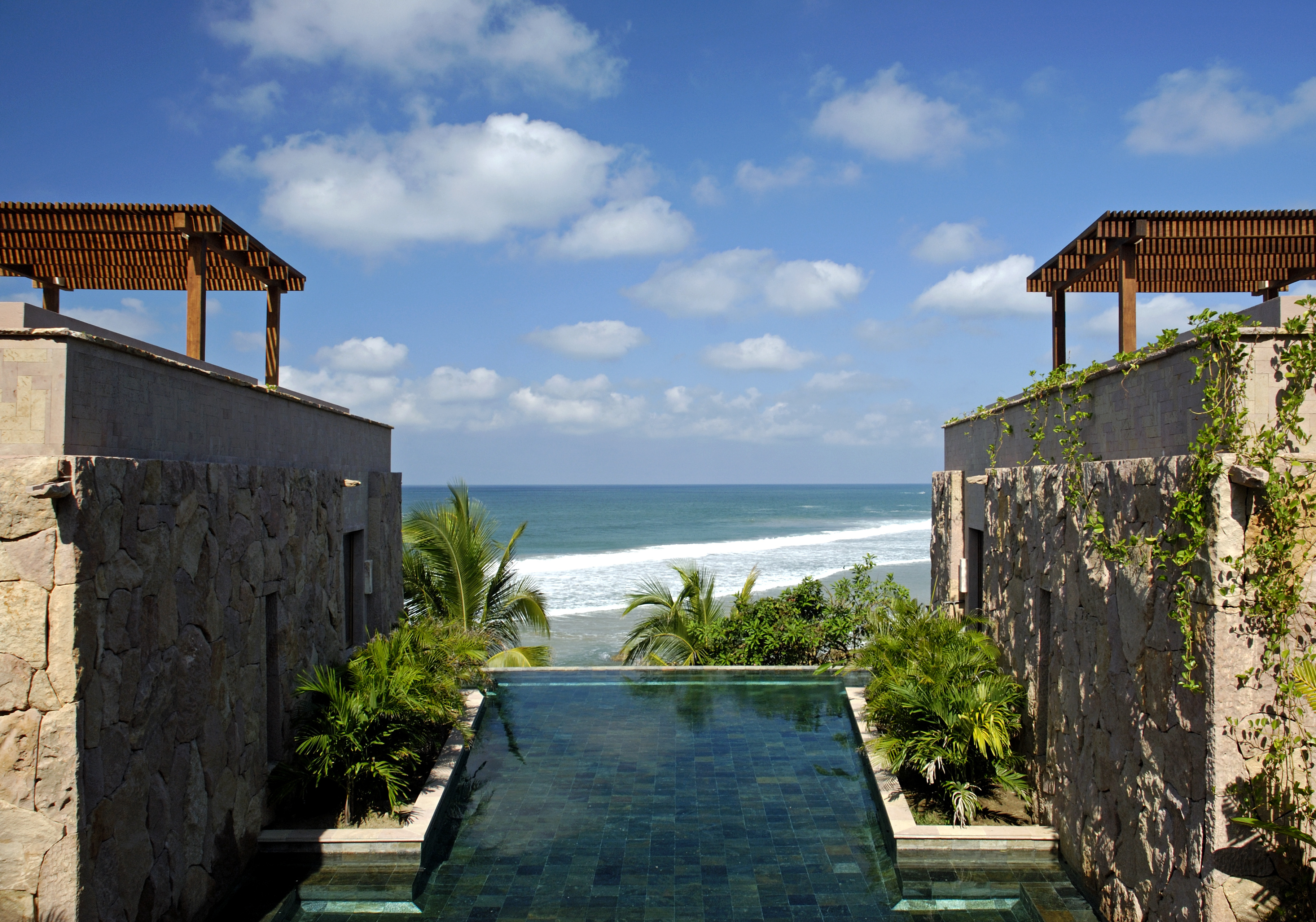 Infinity pool with a view of the ocean, flanked by stone buildings and surrounded by tropical plants under a partly cloudy sky at the luxurious Imanta Resort Spa.