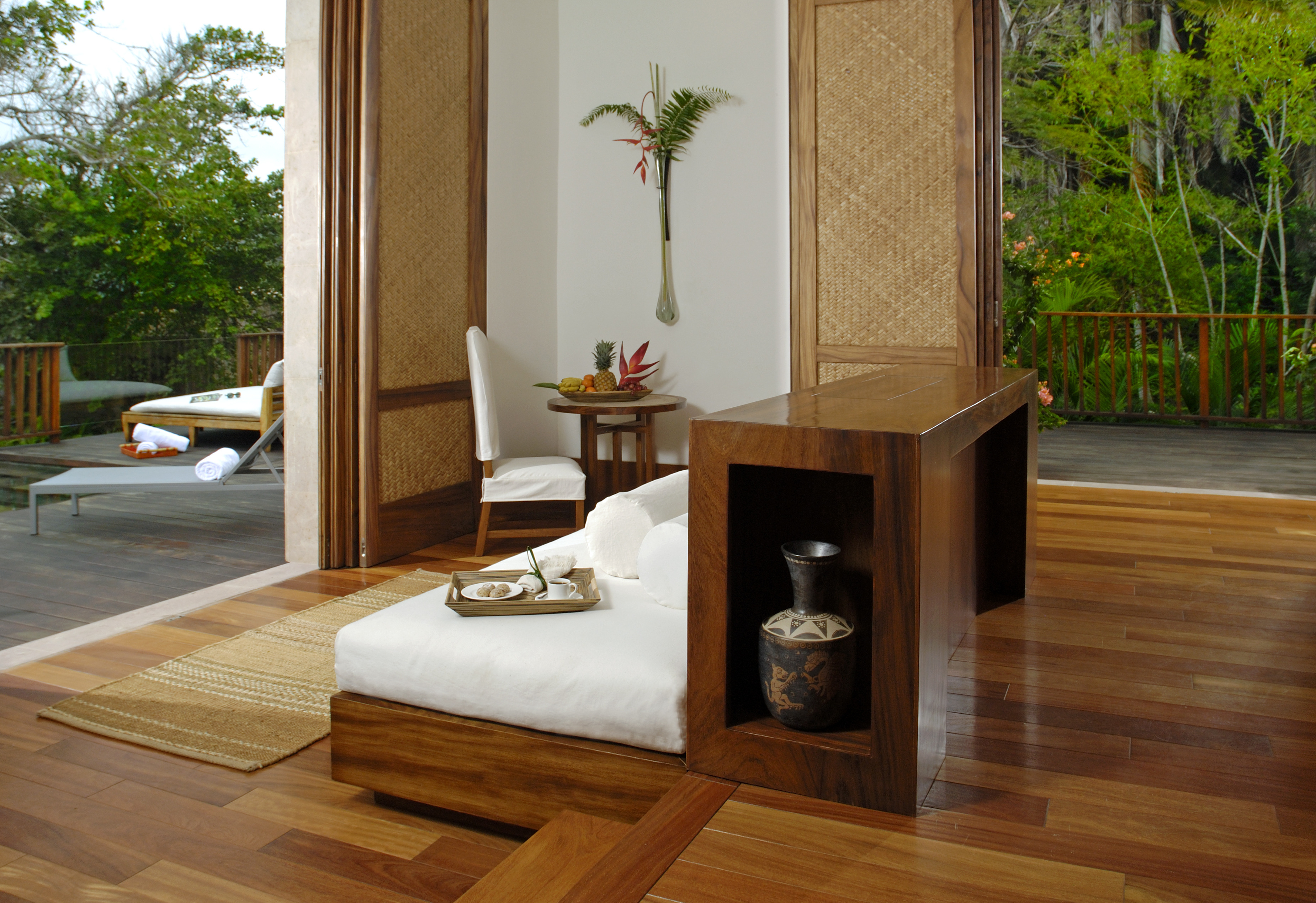 A modern interior features a wooden lounge chair with a tray of dishes, a small table with a chair, and a vase. The space, reminiscent of an Imanta resort spa, opens to an outdoor deck with seating.